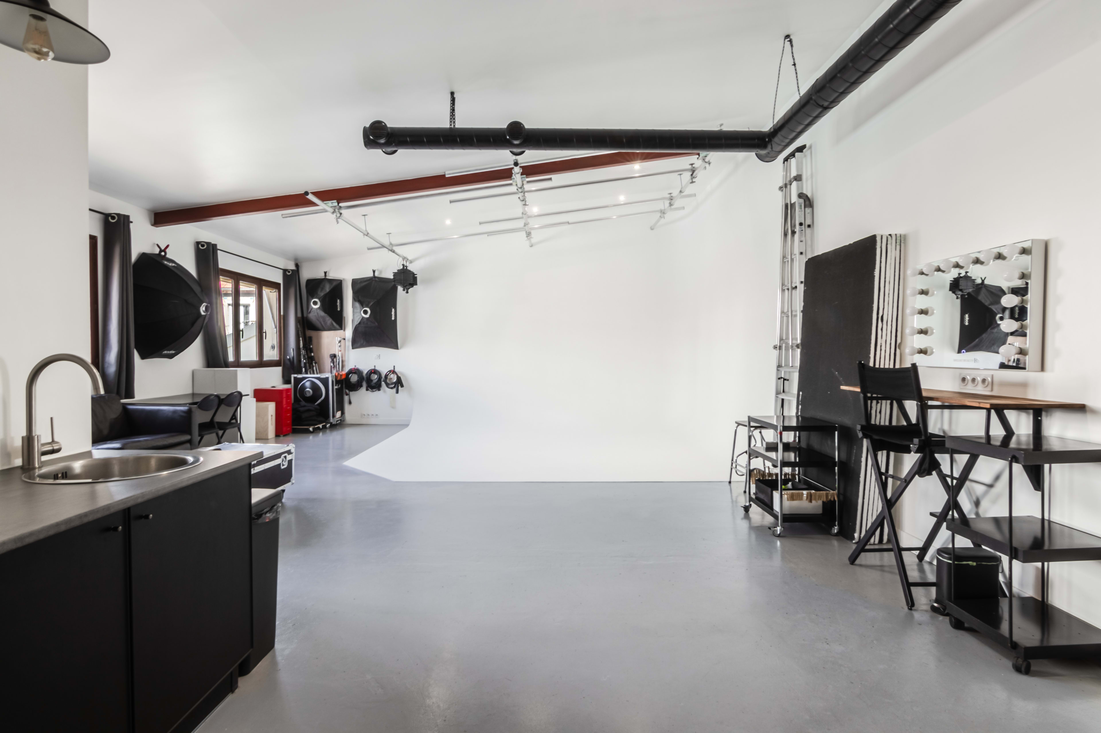 A spacious photography studio features a white backdrop, lighting equipment, a kitchenette area, and a set of black folding chairs along with a mirror.