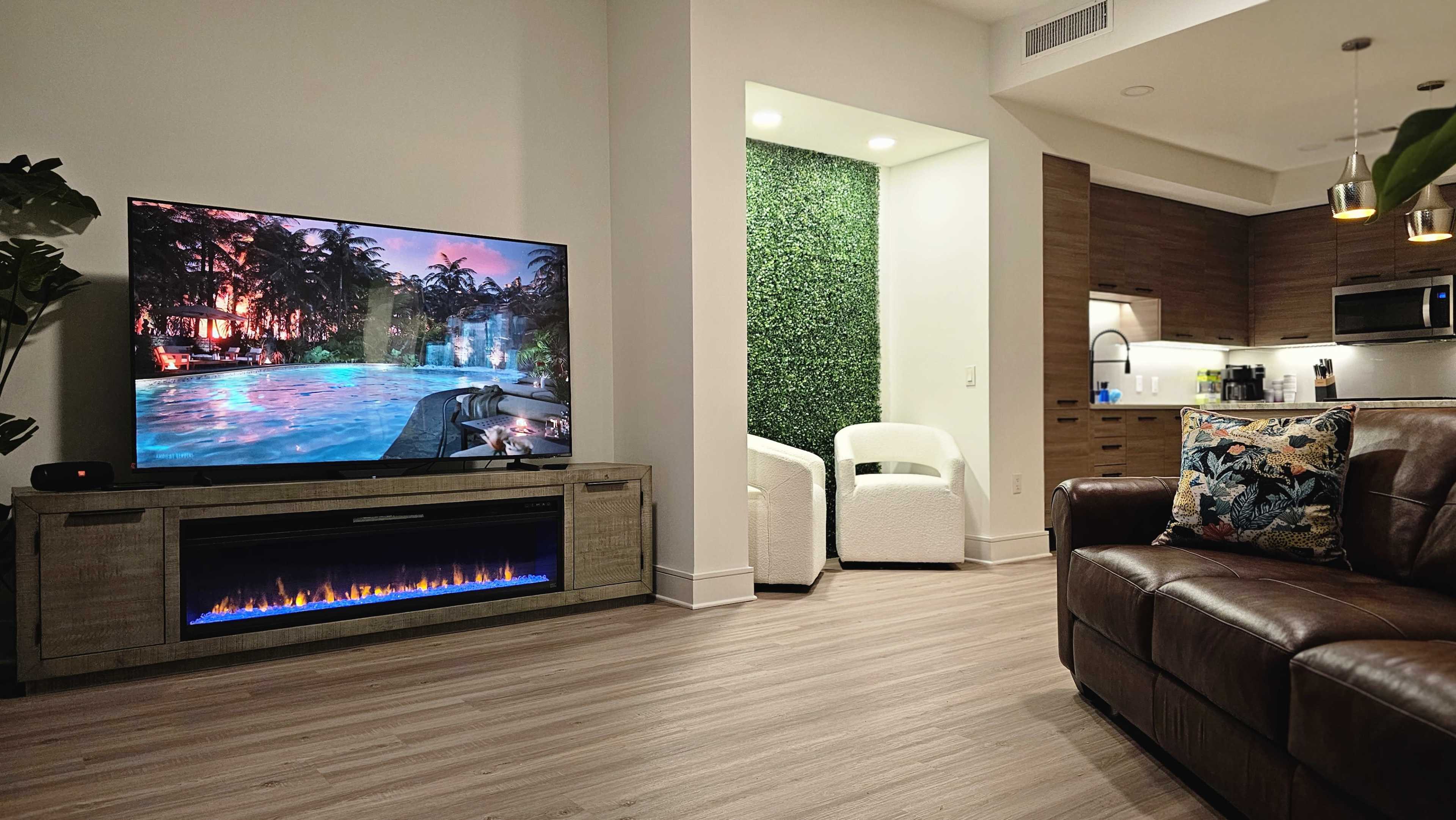 A modern living room featuring a large television displaying a pool scene, a sectional sofa, and a small kitchenette area with light wood cabinetry in the background.