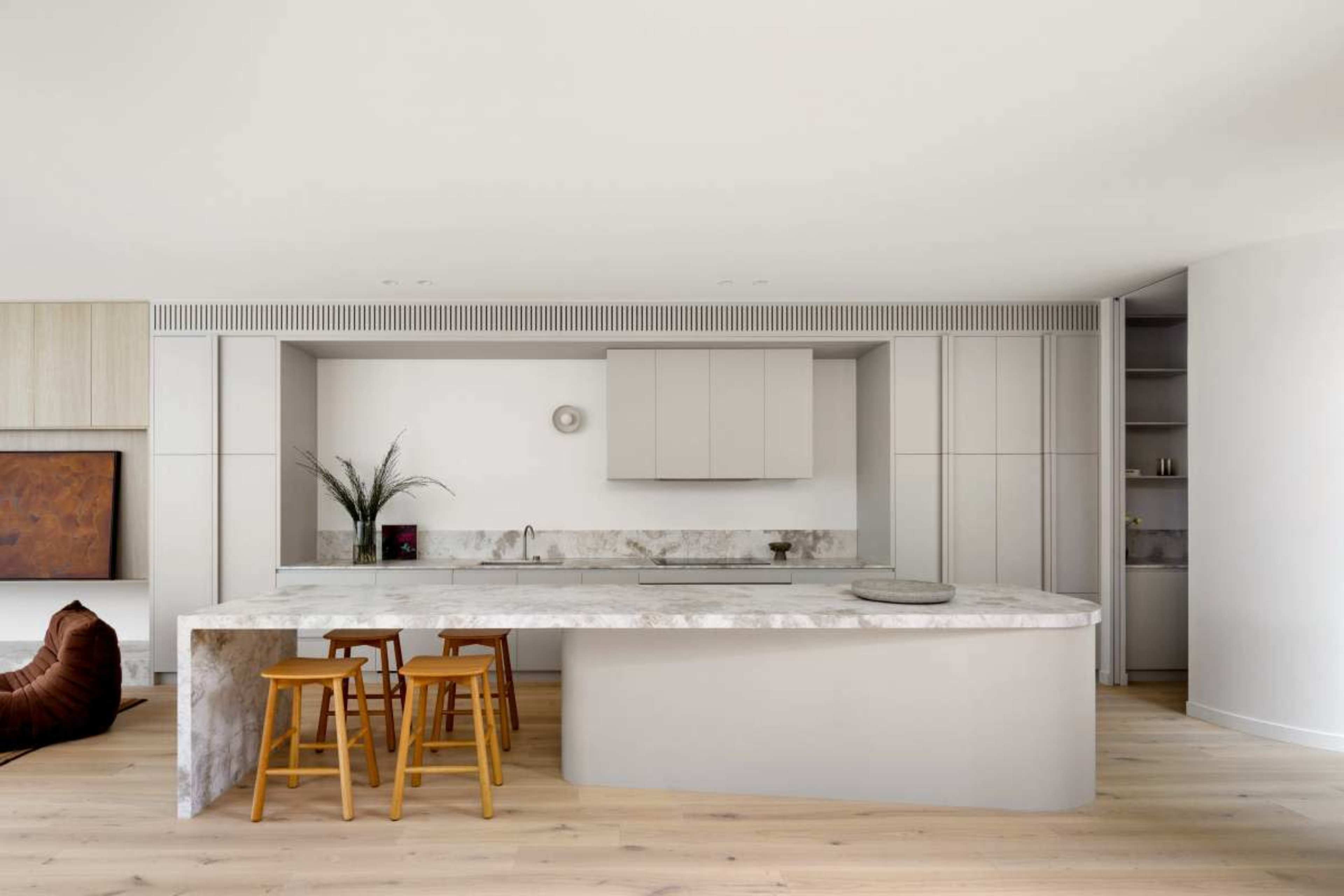 The image shows a modern kitchen with a large marble island and light wood stools, featuring sleek cabinetry and a minimalist design.