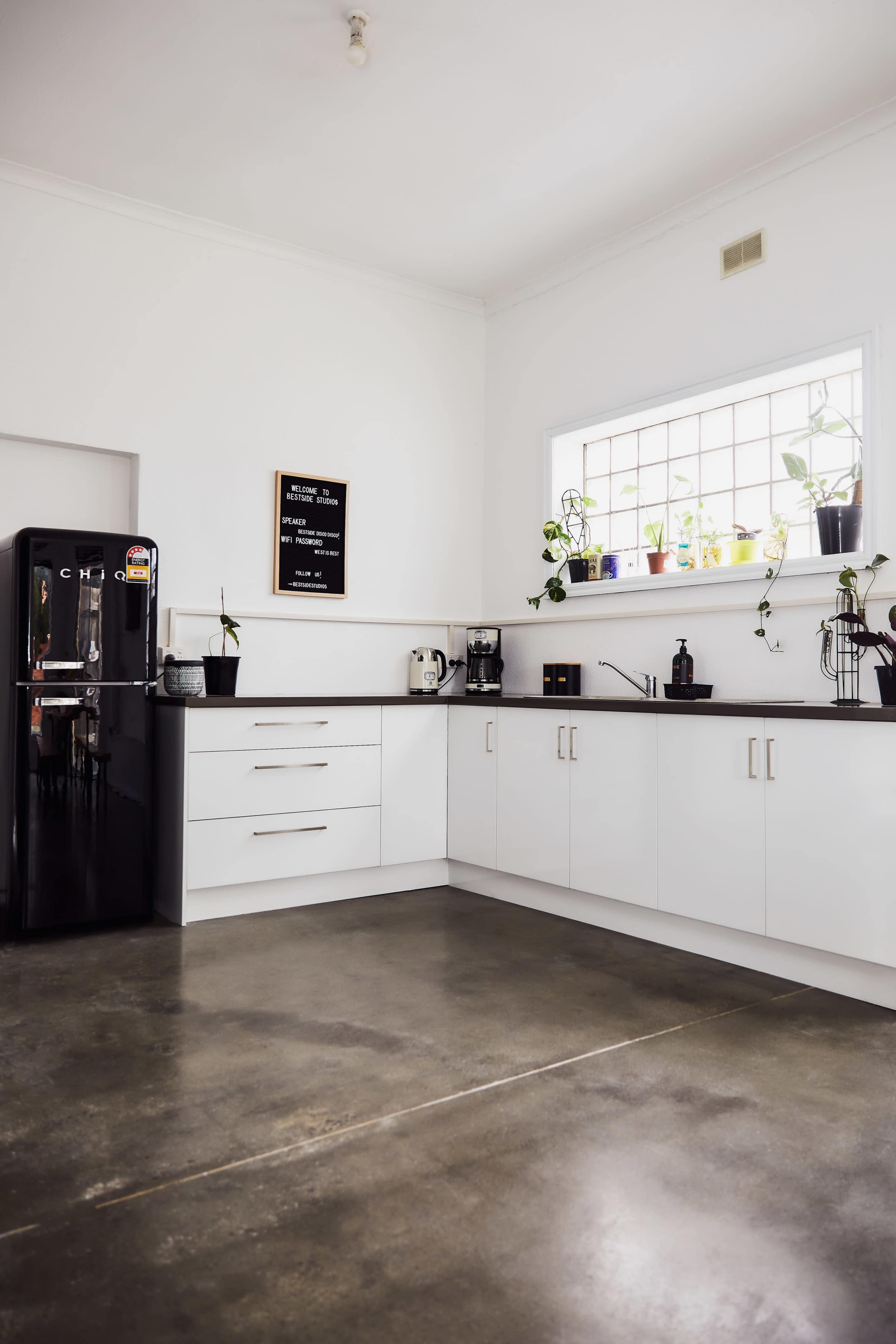 A modern kitchen features a black refrigerator, white cabinetry, and a window lined with potted plants.