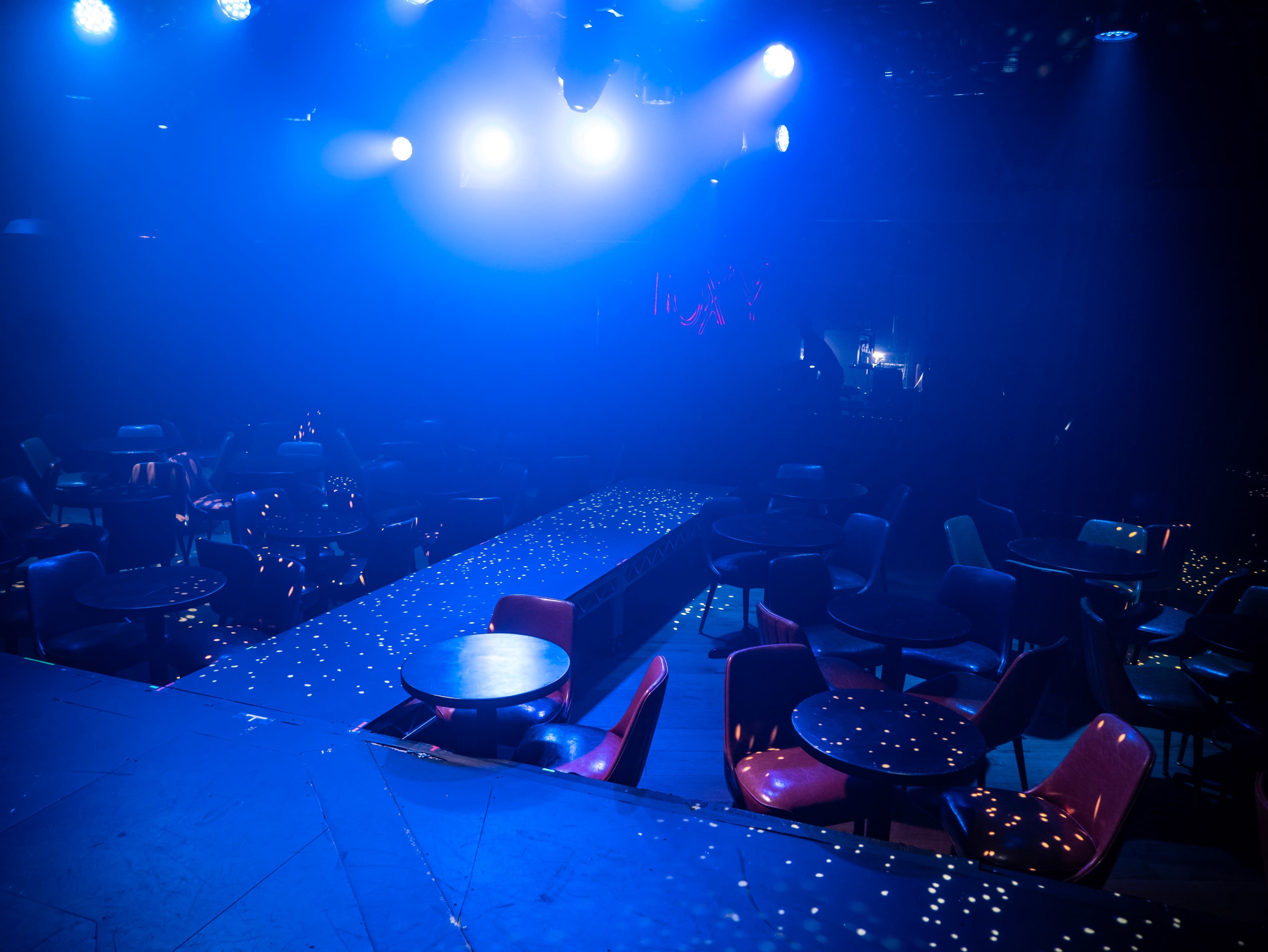A dimly lit nightclub features scattered tables and a runway illuminated by blue lights.