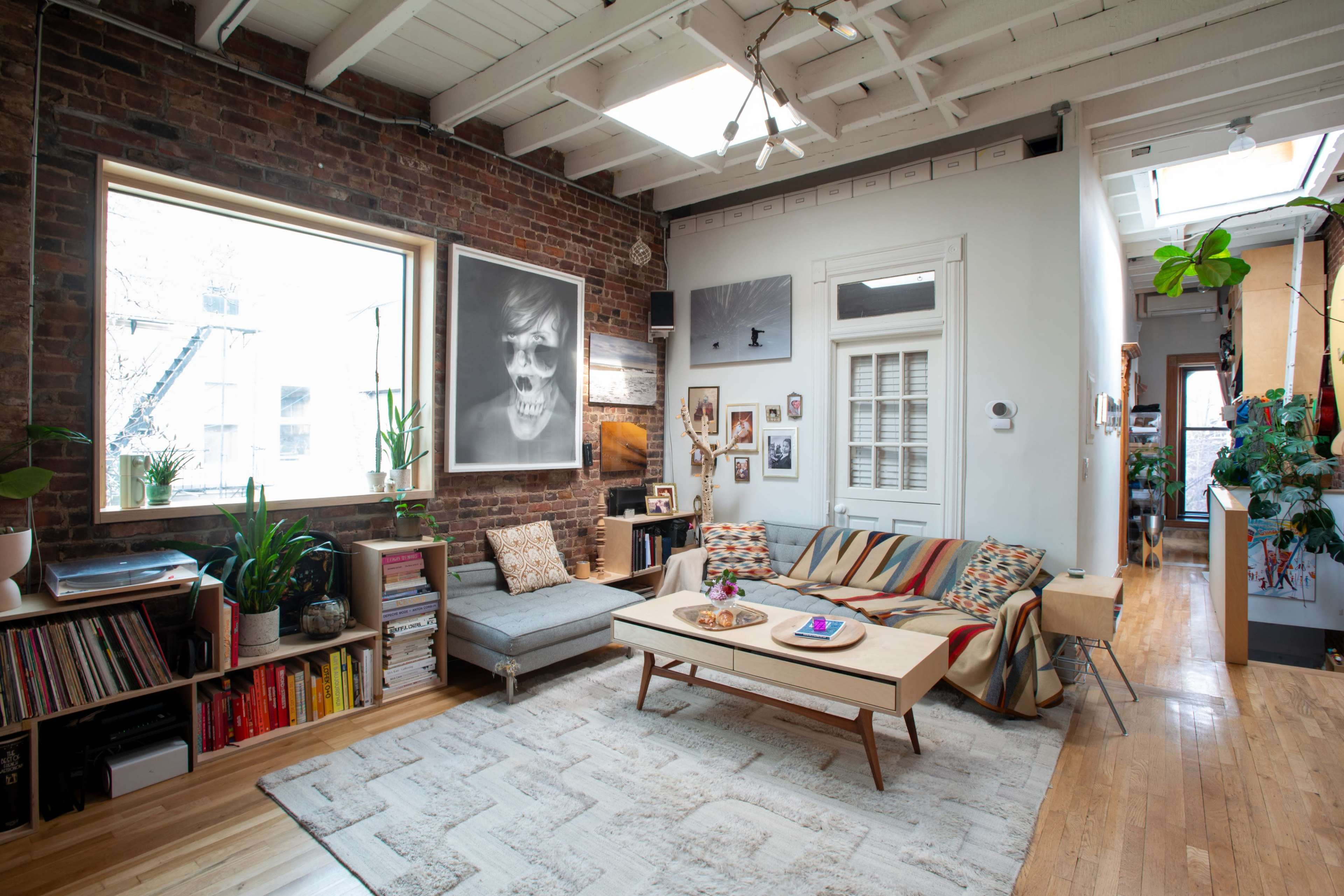 The image shows a cozy living room featuring exposed brick walls, large windows, a mix of seating options, and various plants.