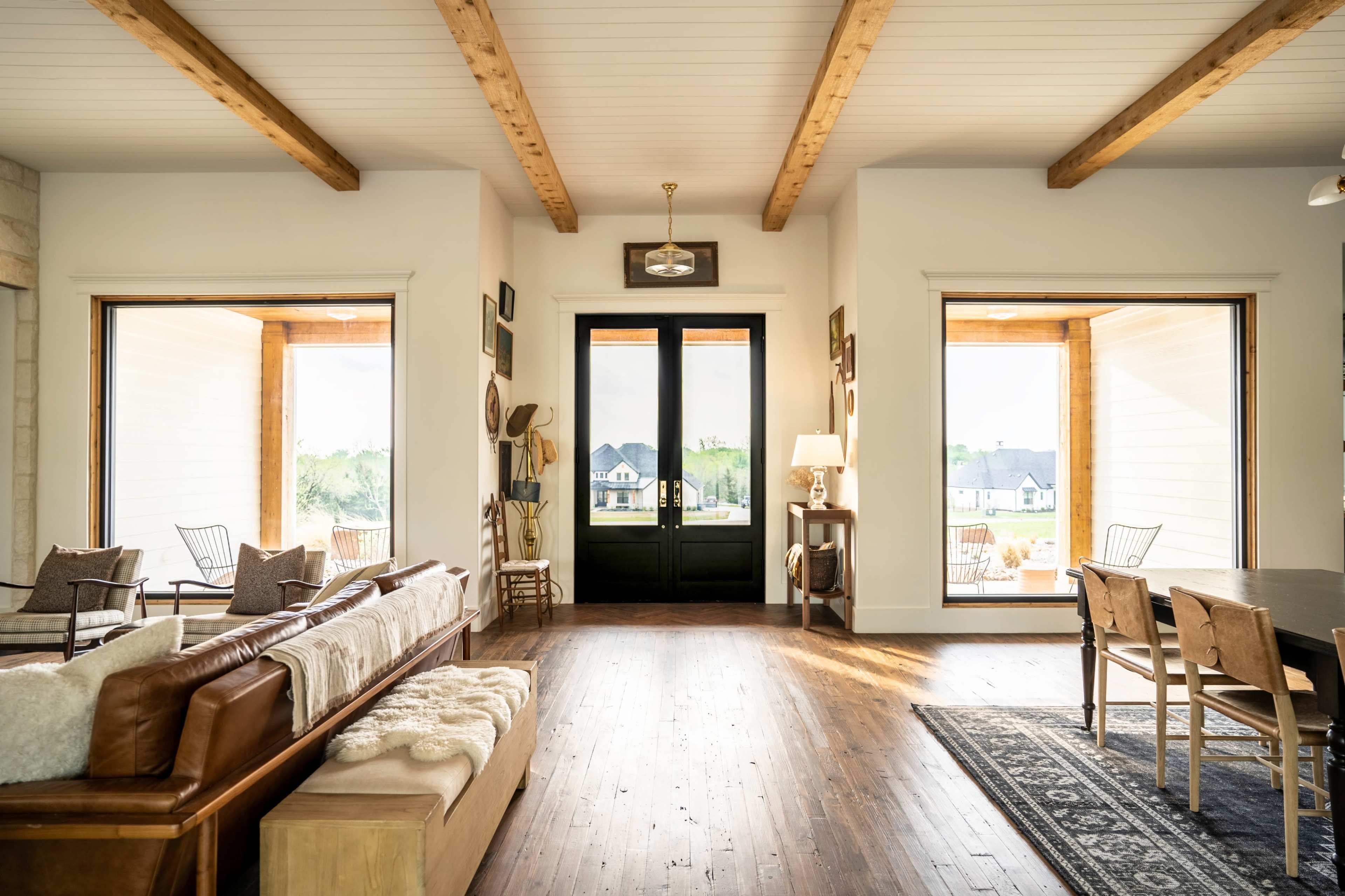 A spacious interior features large windows that frame a view of the outdoors, with wooden beams overhead and minimalist furnishings throughout.