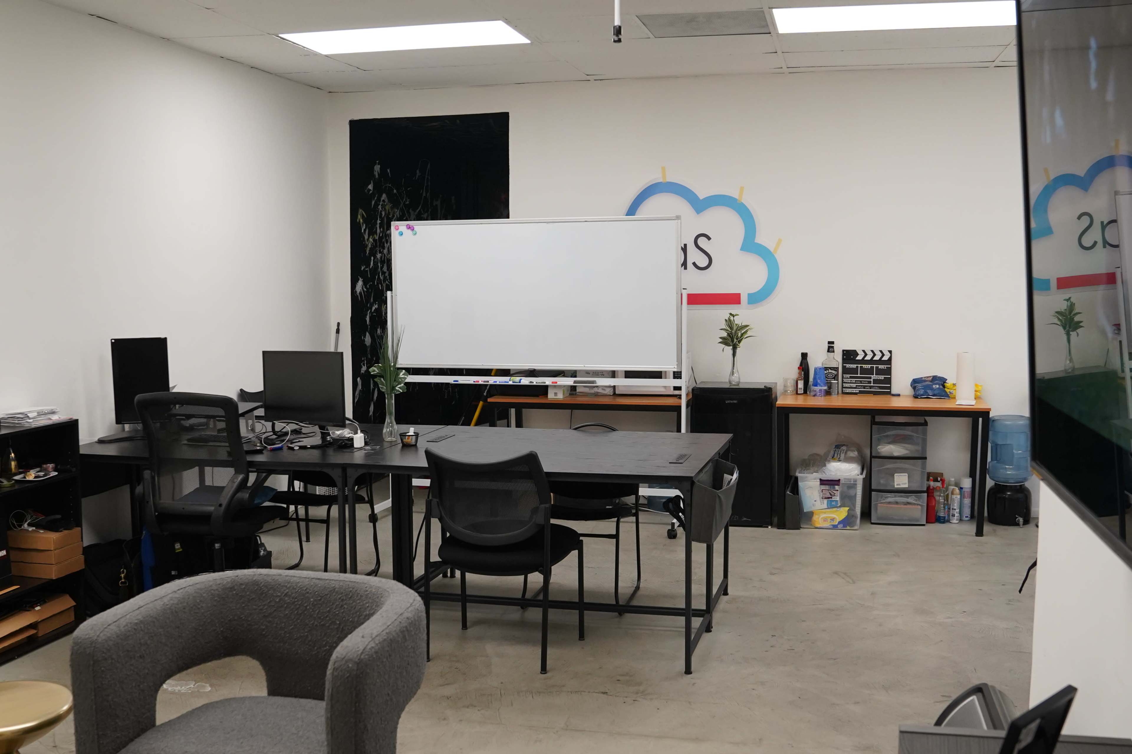 The image shows an empty office space featuring a large table with chairs, a whiteboard on a stand, and workstations along the walls, with shelves and a water cooler in the background.