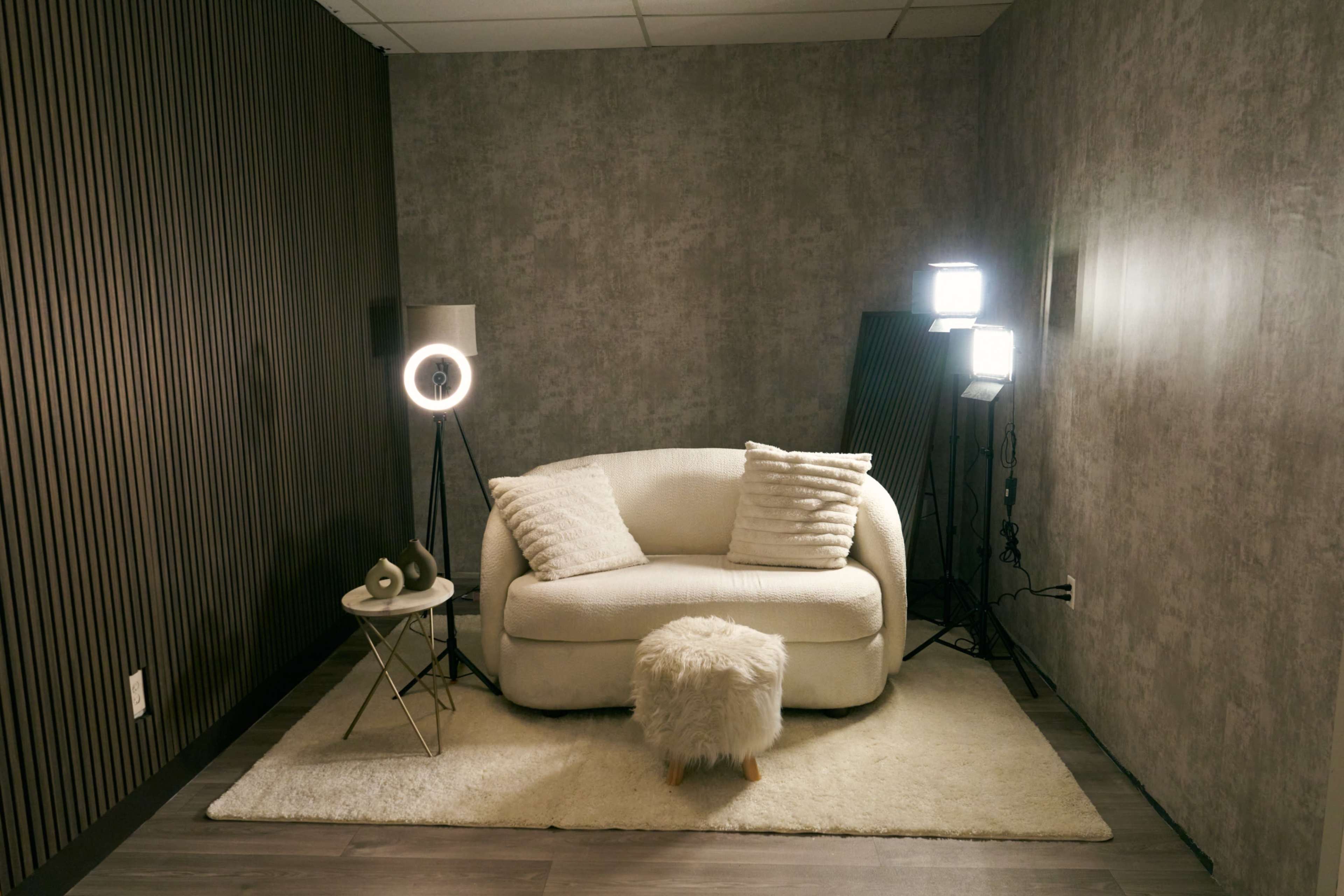 The image shows a cozy corner featuring a white sofa with cushions, a small round side table, a fluffy ottoman, and lighting fixtures against a textured wall.