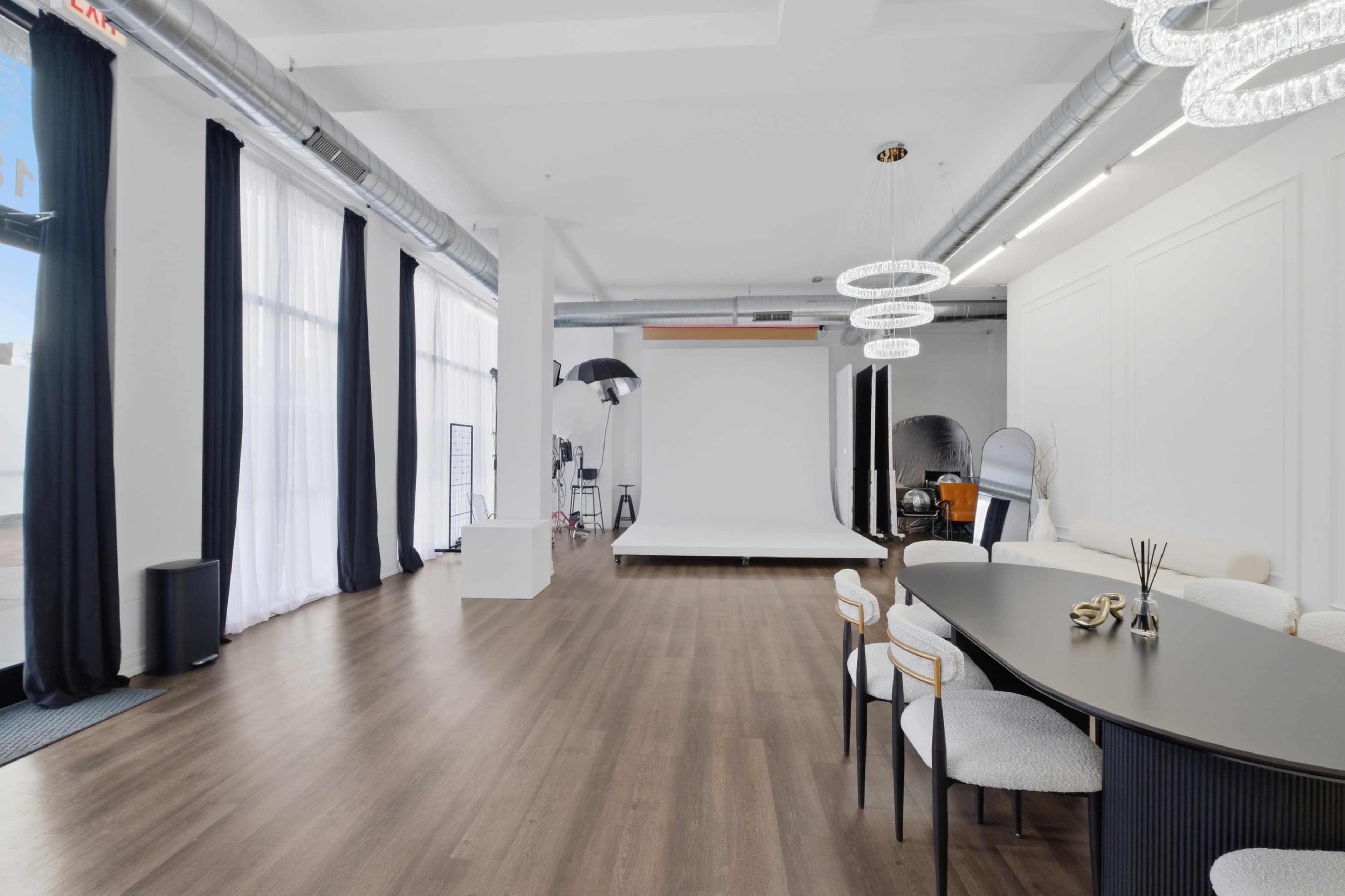 The image shows a spacious, minimalist studio with large windows, a white backdrop, and a dining table with chairs to one side.