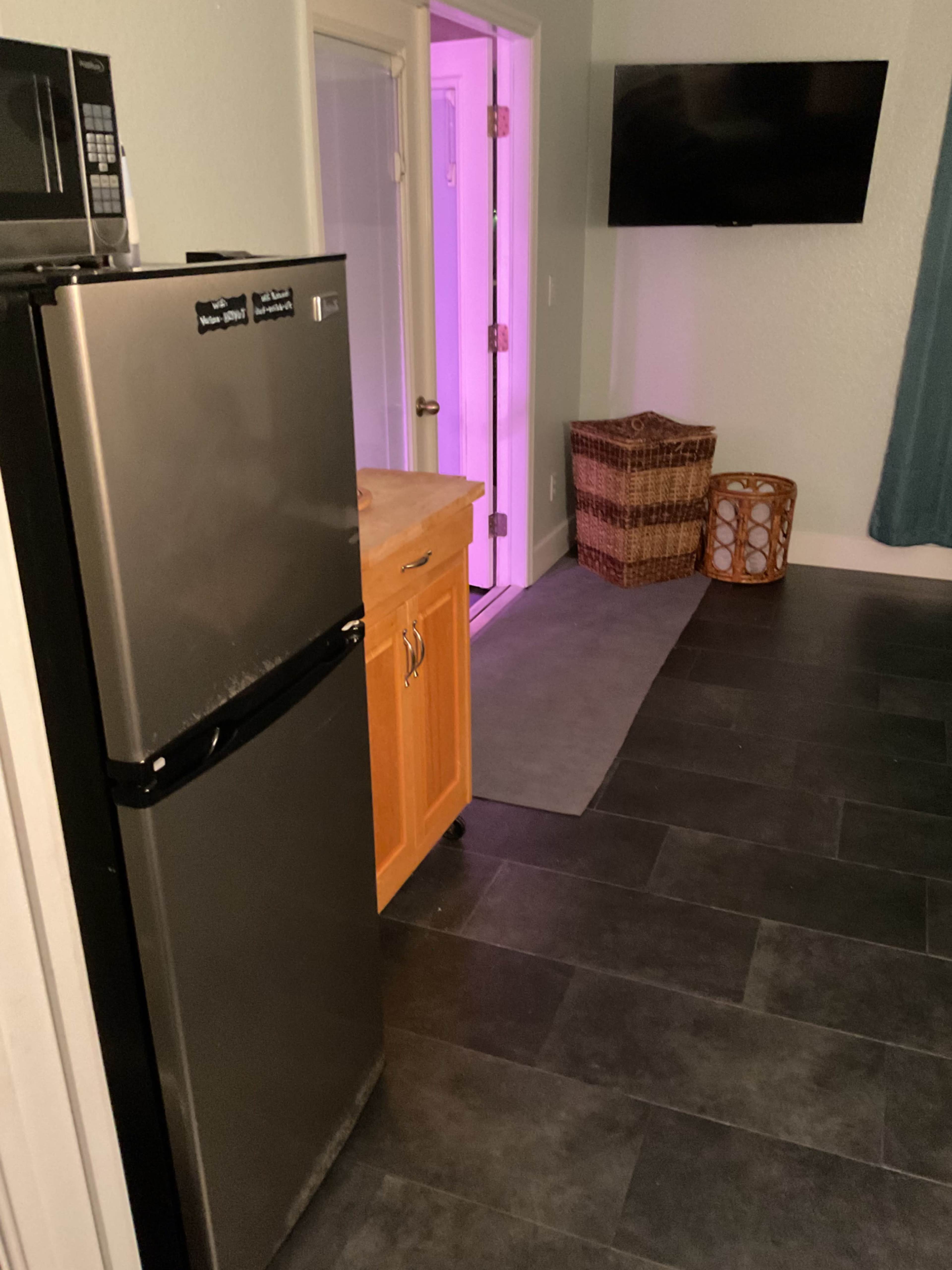 The image shows a small kitchen area with a stainless steel refrigerator, a wooden countertop, a woven basket, and a visible doorway illuminated by purple light.