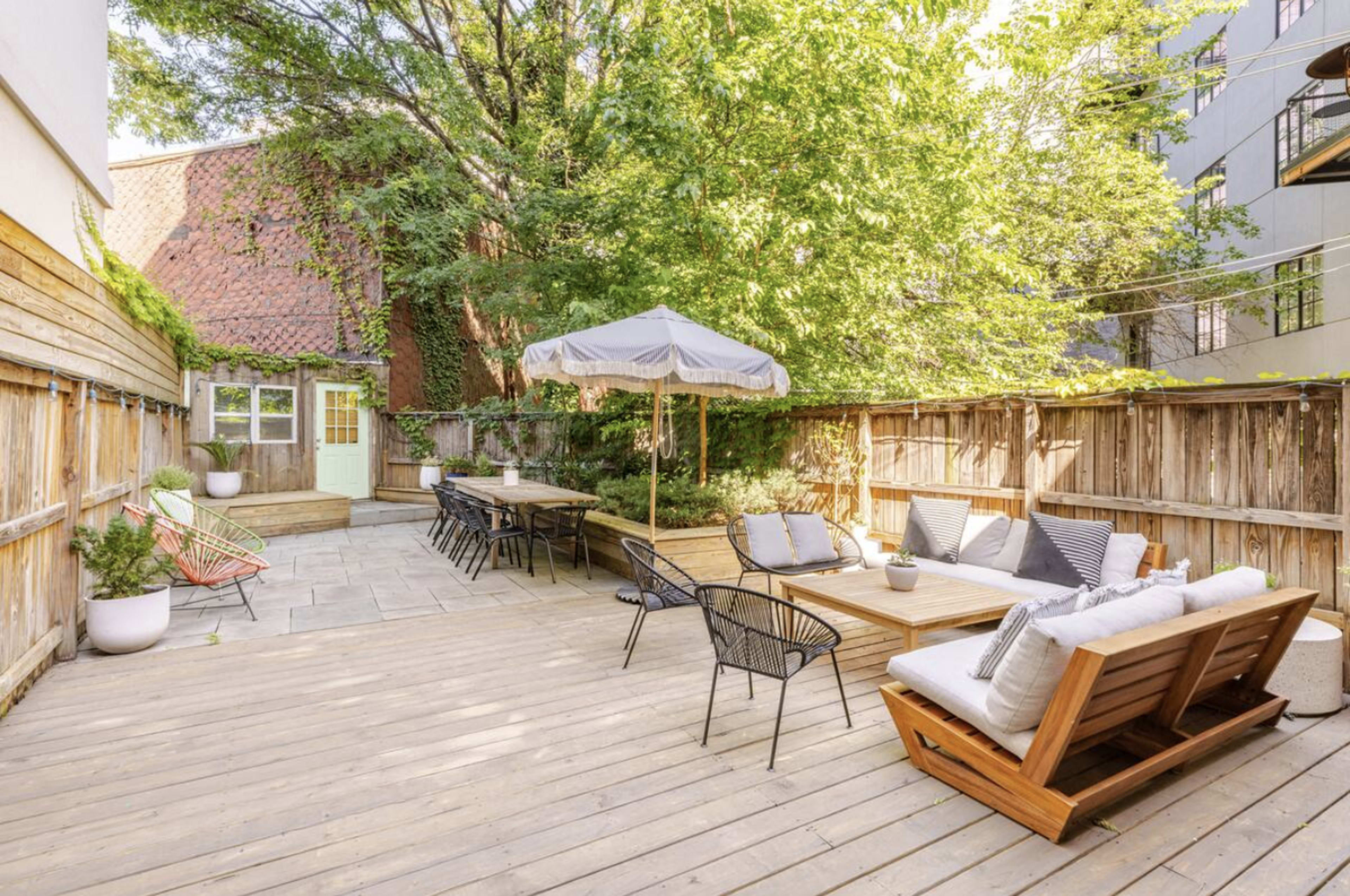 The image shows a wooden deck area furnished with outdoor seating, a large umbrella, and a dining table, surrounded by greenery and a wooden fence.