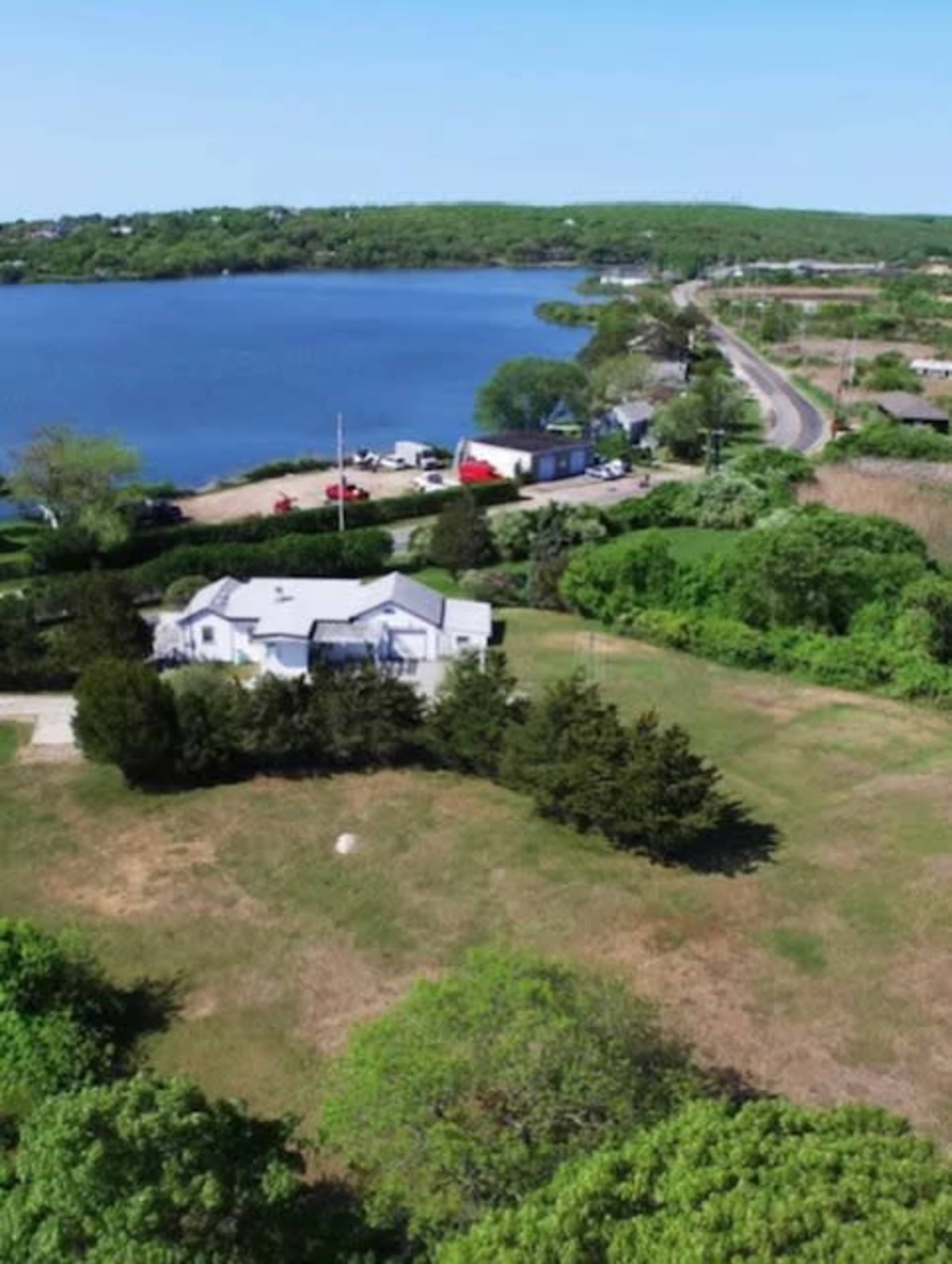 A lake bordered by green trees and a road with several parked vehicles alongside a house in the foreground.