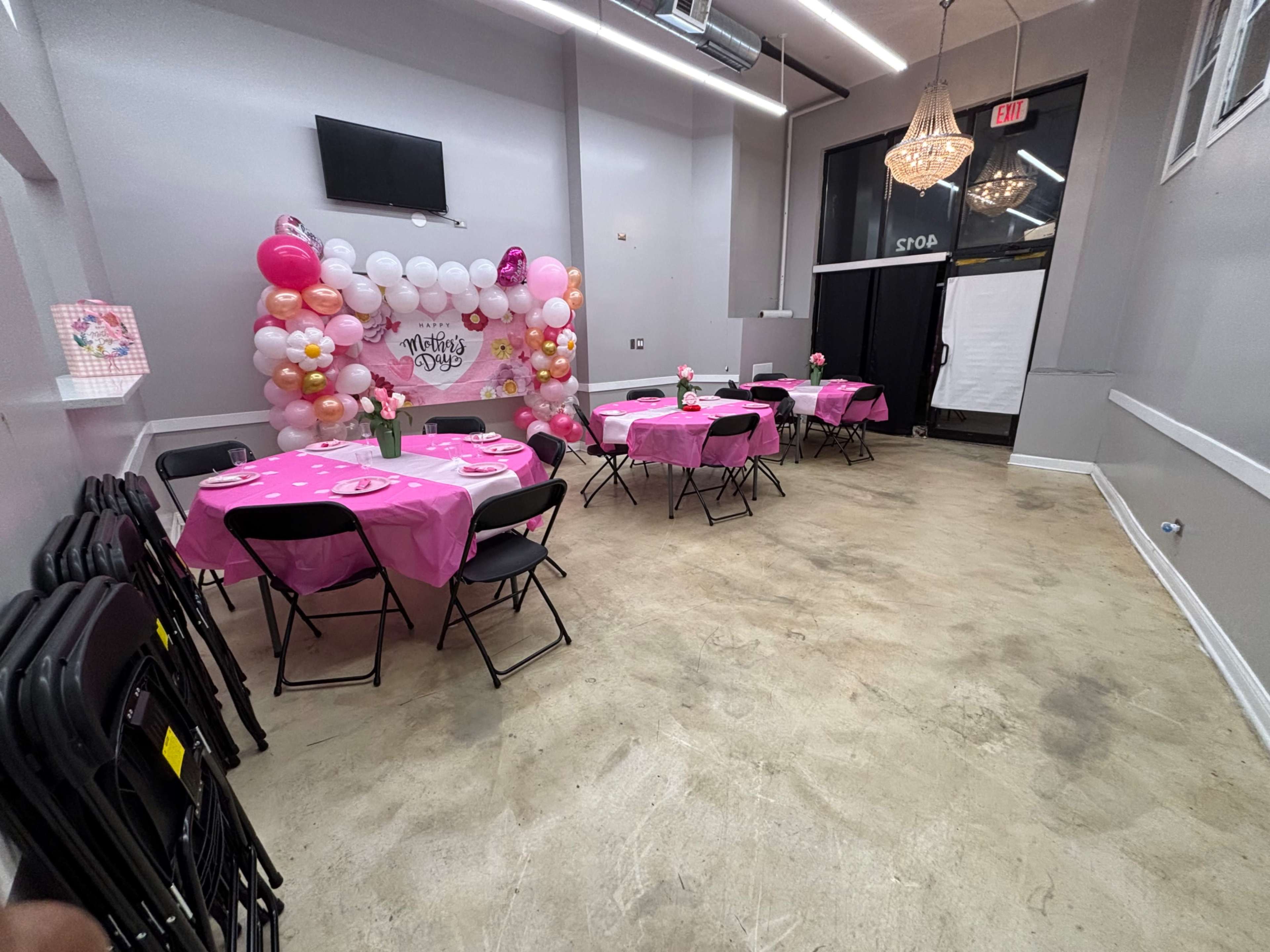 The image shows a decorated event space with pink tablecloths, black chairs, and a balloon backdrop for a Mother's Day celebration.