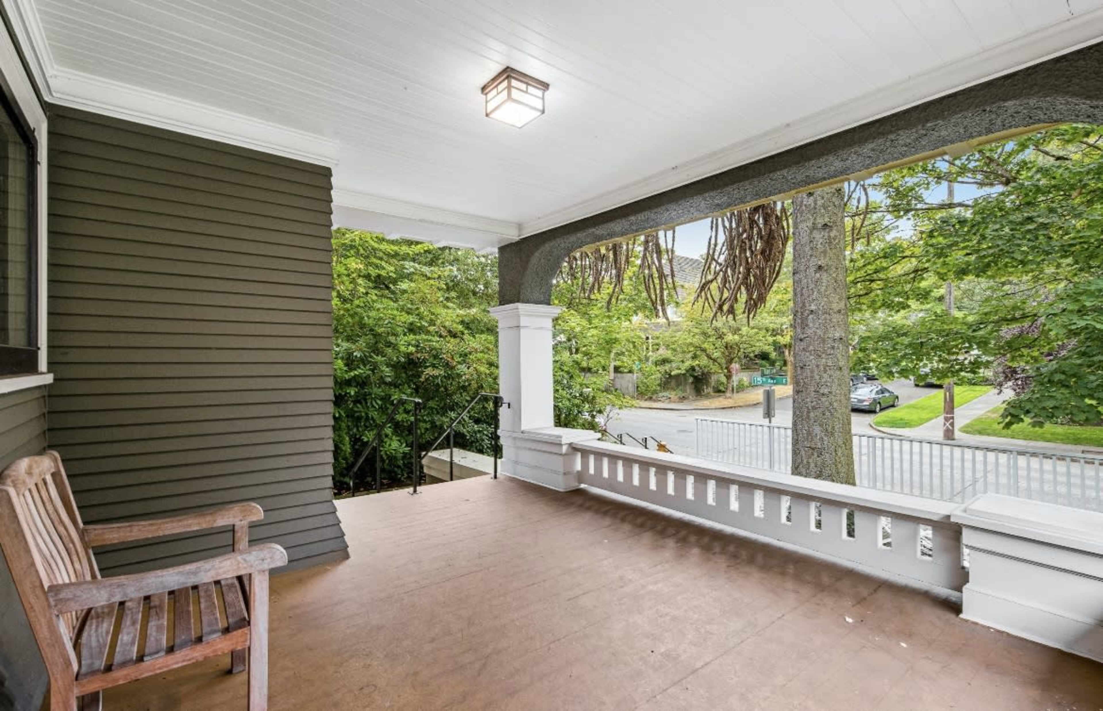 A covered porch with a wooden bench, surrounded by greenery and a view of a street.