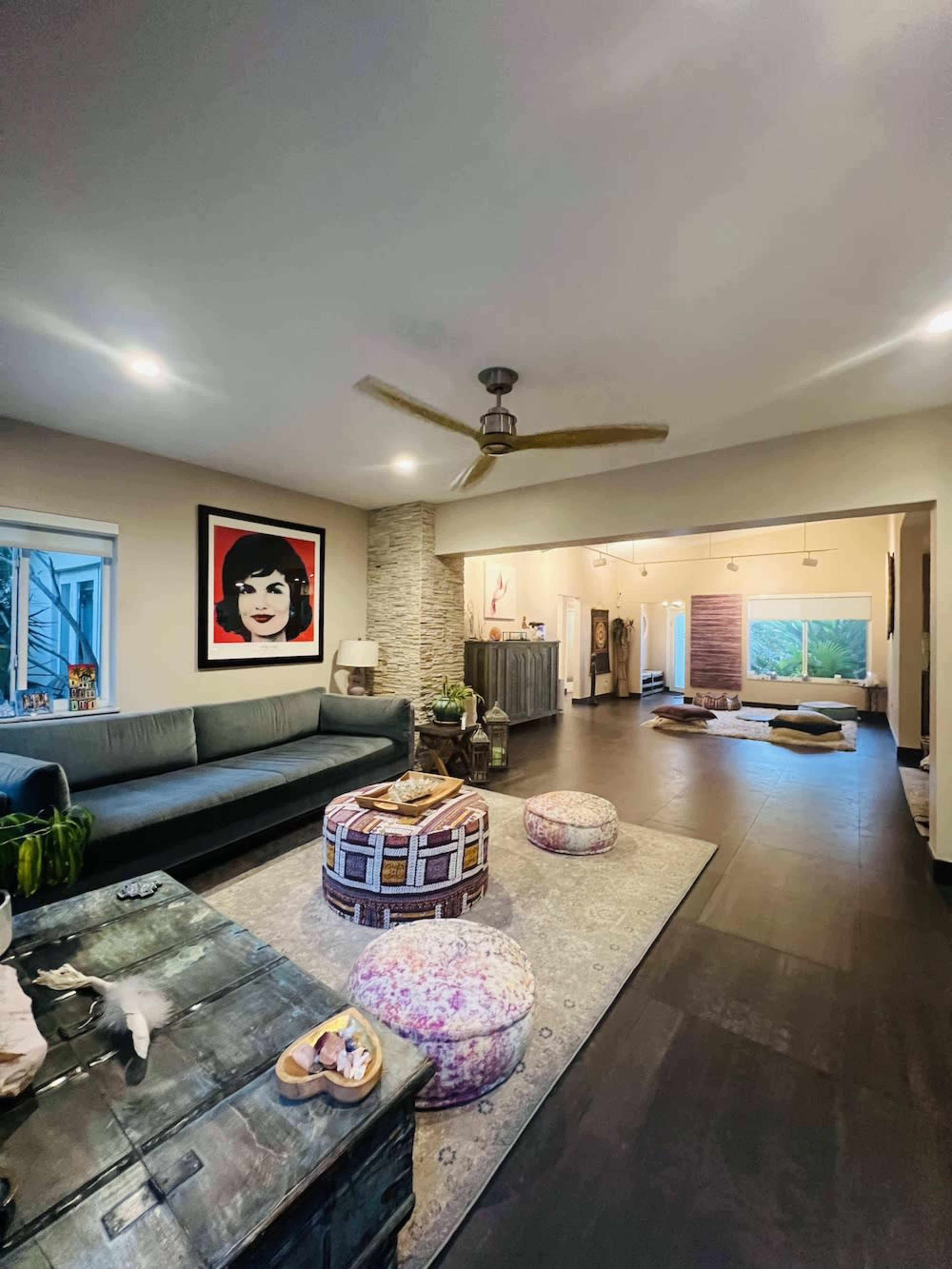 A spacious living room with a large sofa, a decorative rug, and a prominent artwork on the wall, leading into a well-lit area with additional seating and windows.