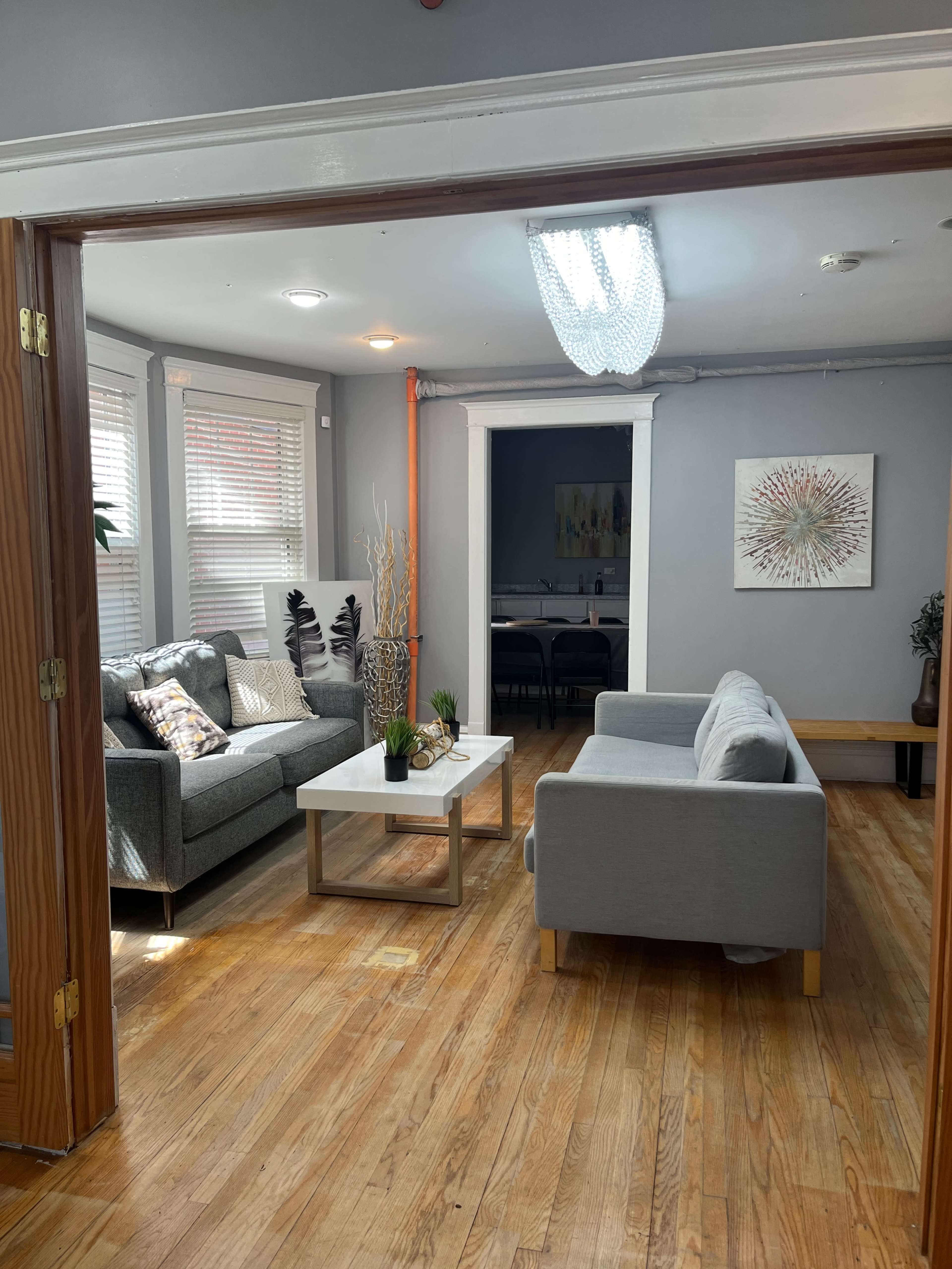 The image shows a living area with two gray sofas, a coffee table, and decorative plants, featuring wooden flooring and natural light from windows.