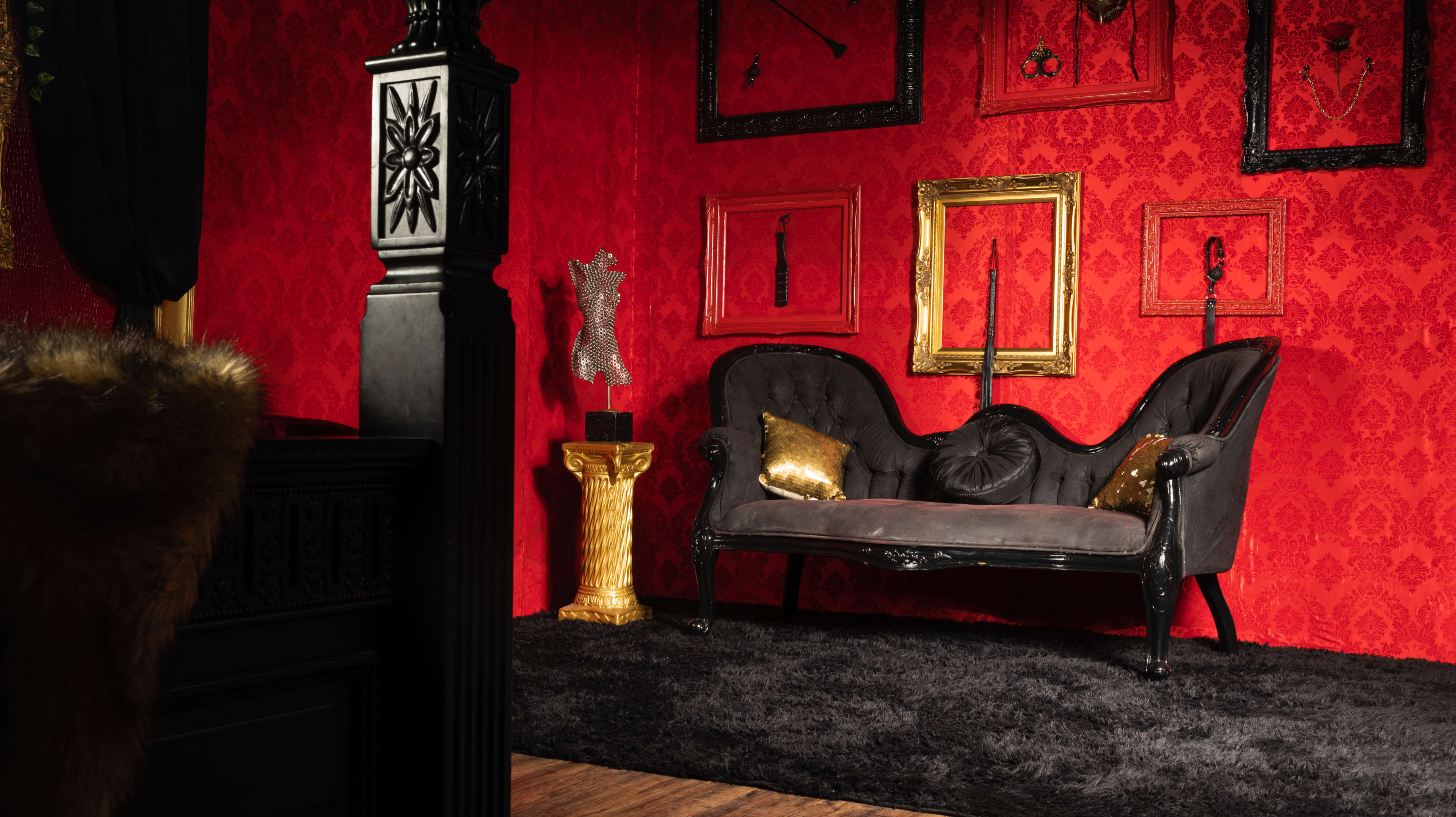 A dark Victorian-style sofa against a bold red patterned wall adorned with various ornate frames.