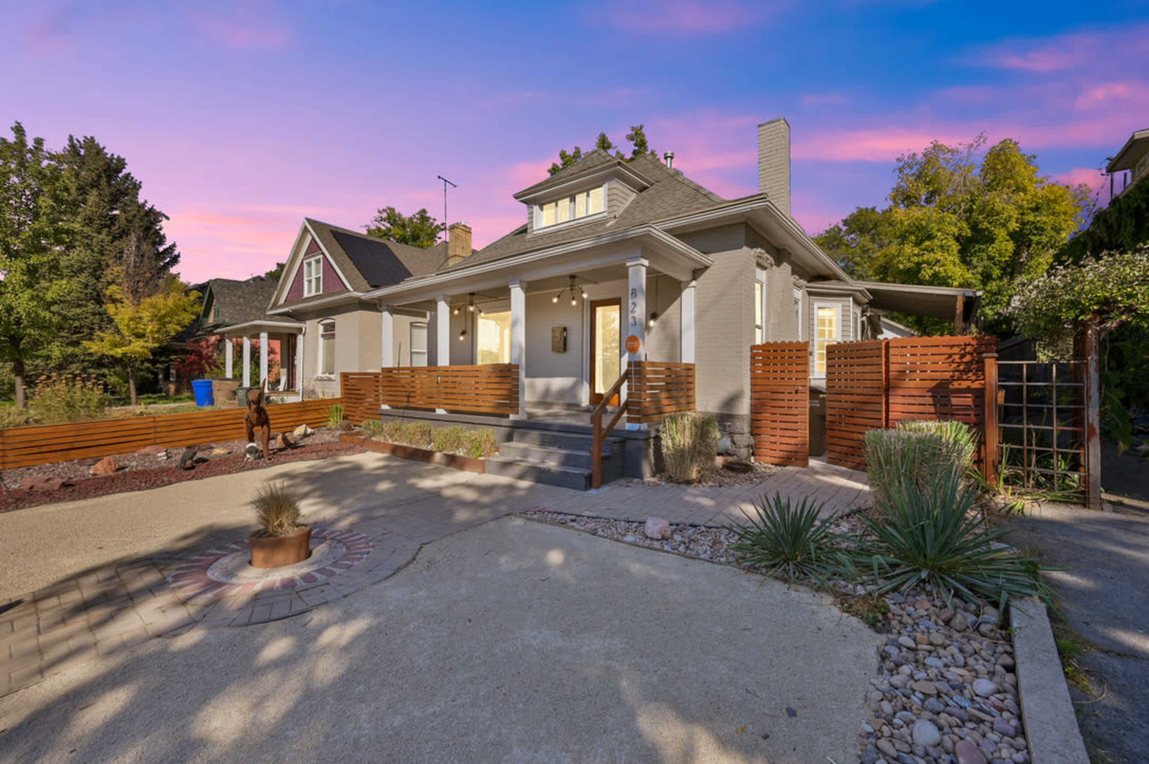 A gray single-story house with a front porch, surrounded by a landscaped yard and wooden fencing, set against a colorful sky at dusk.