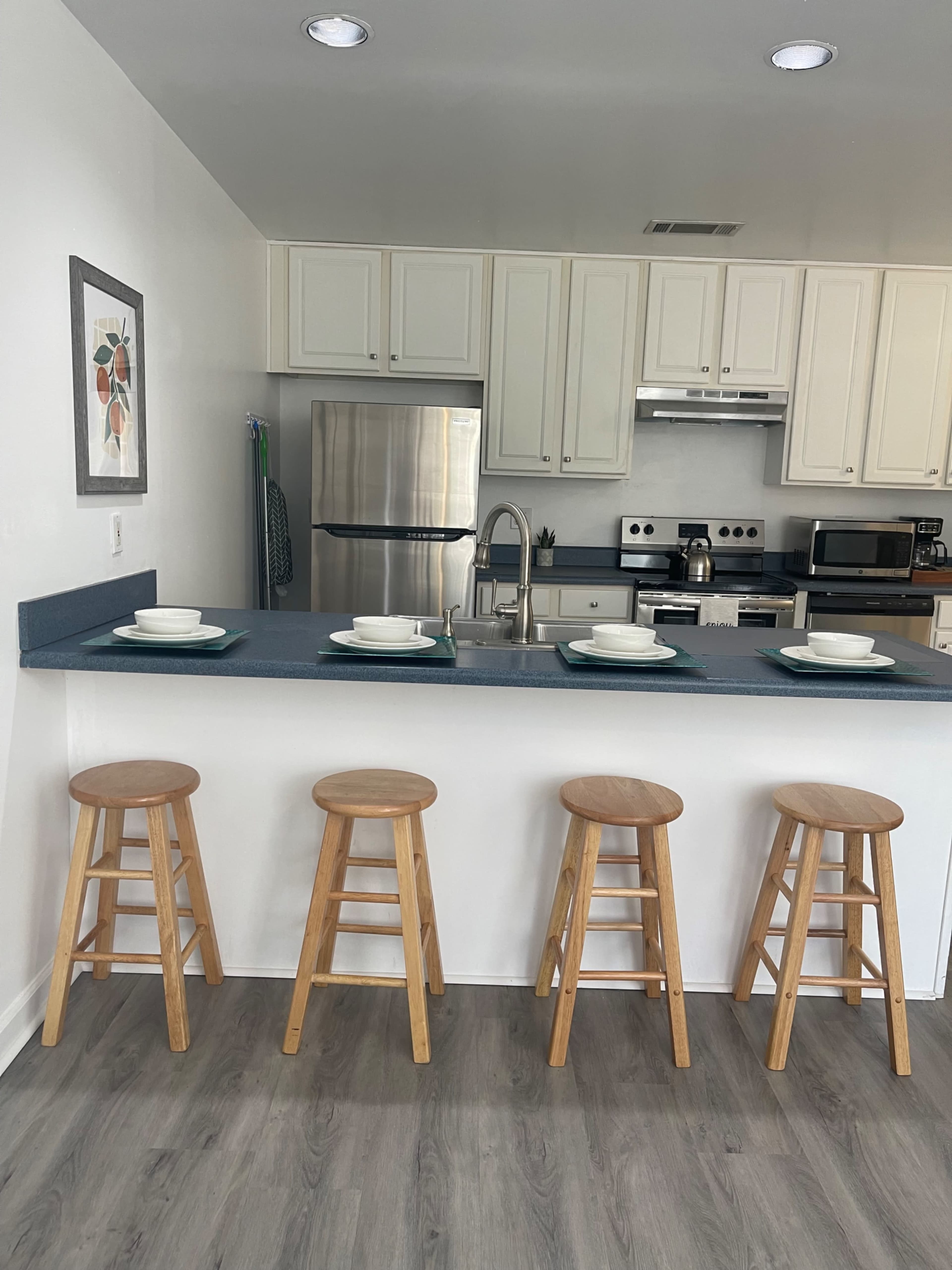 A modern kitchen features a bar with four wooden stools and a countertop set with plates, overlooking stainless steel appliances.