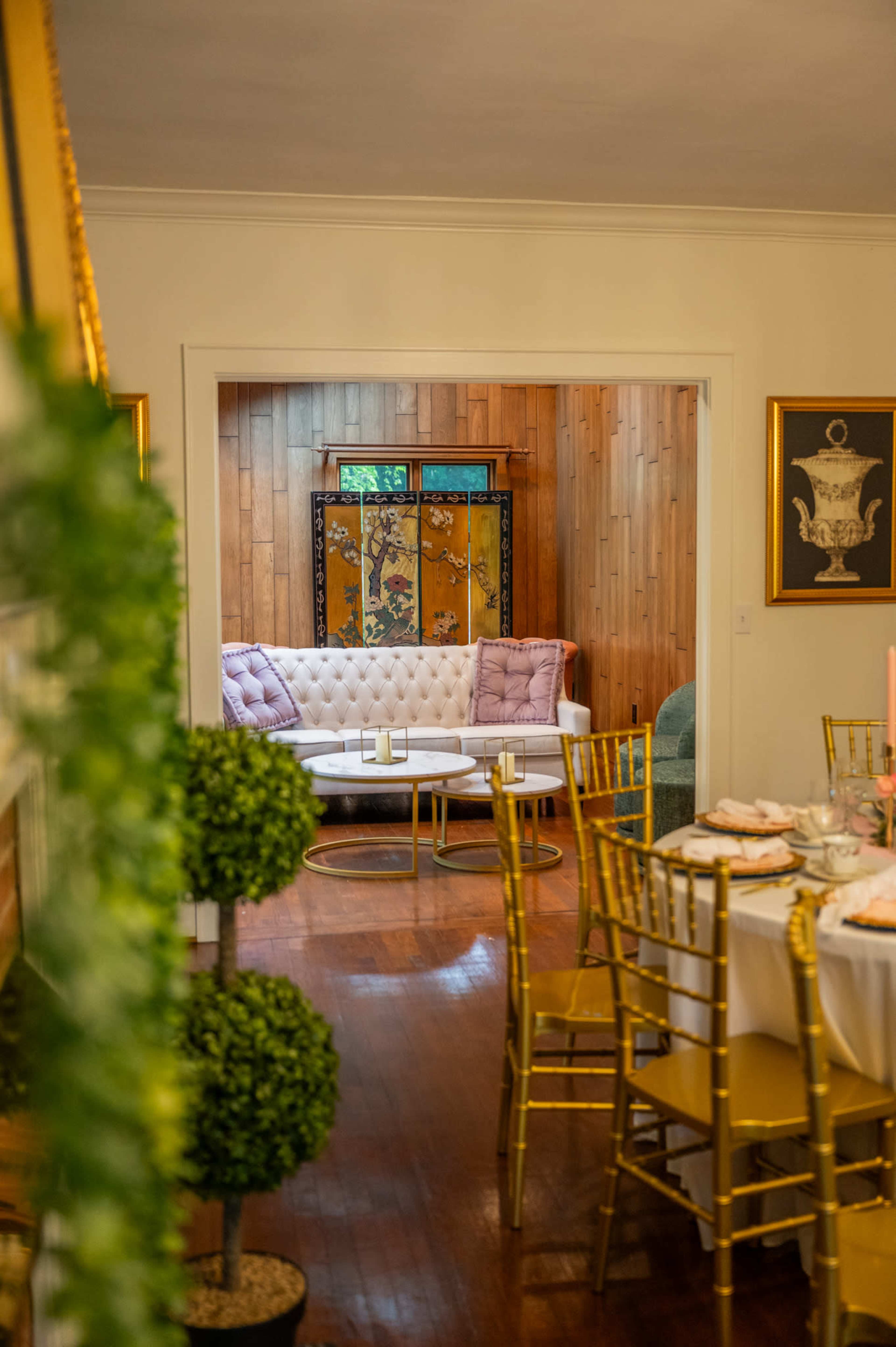 A dining area with gold chairs and a table set for a meal opens to a cozy living space featuring a white sofa and wooden accent wall.