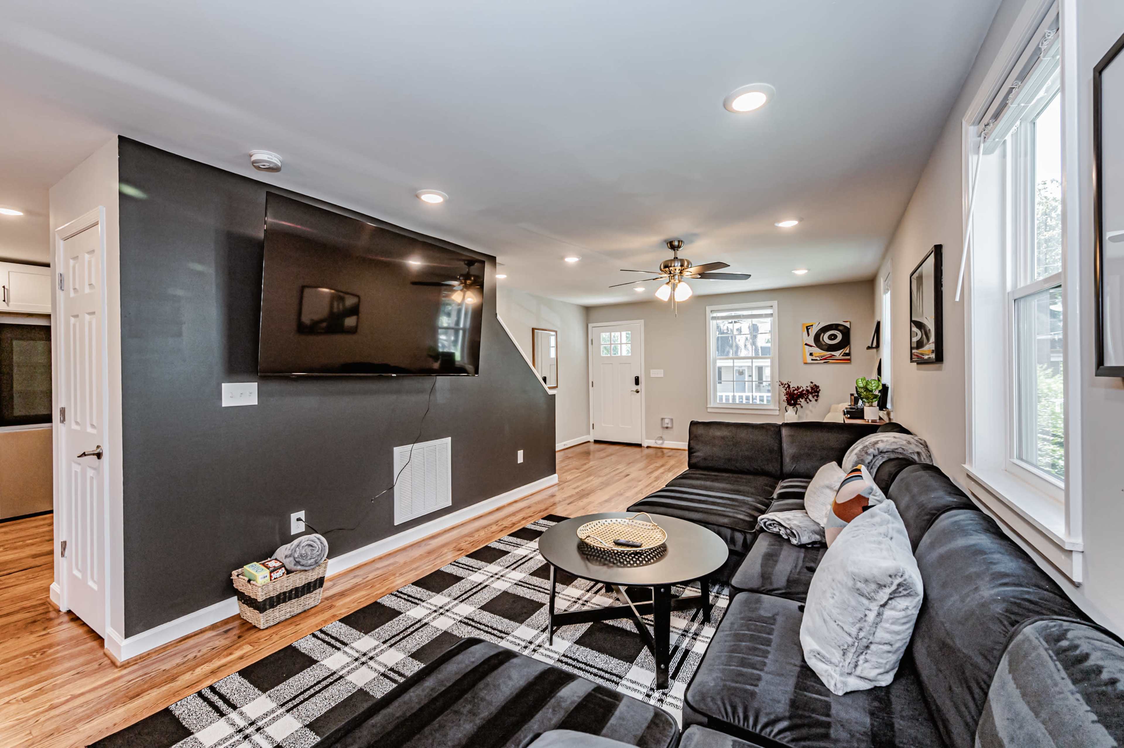 The living room features a large sectional sofa, a coffee table, and a mounted flat-screen TV on the wall, with a view of a staircase leading to another level.