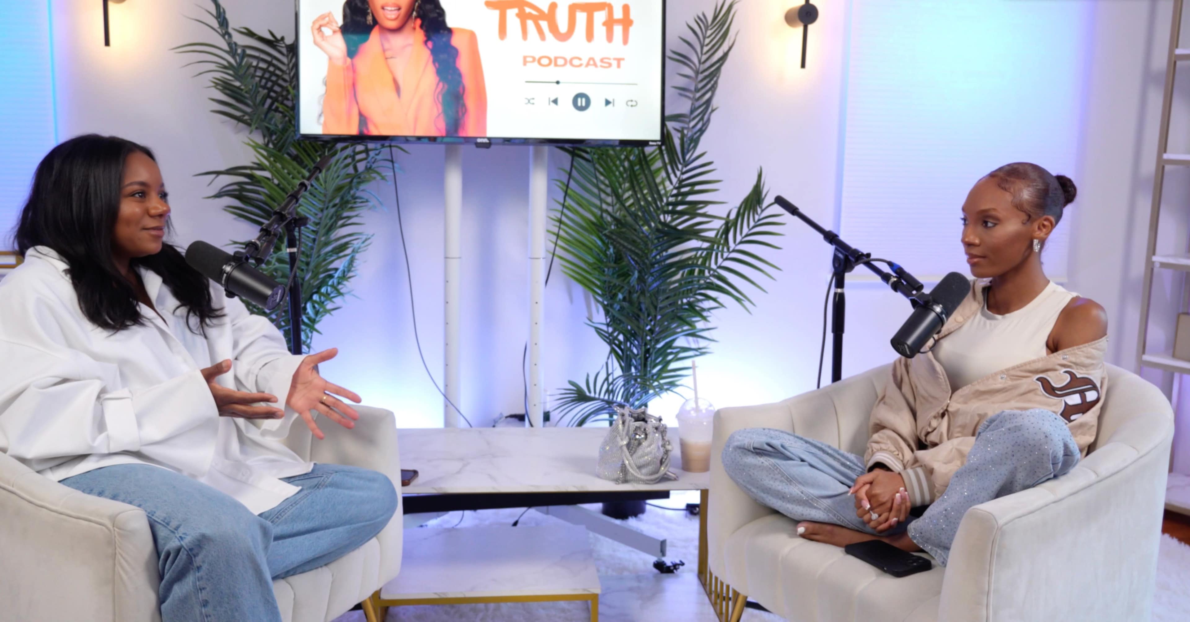 Two women sit in an indoor podcast studio, engaged in conversation, with microphones in front of them and a screen displaying the podcast title in the background.