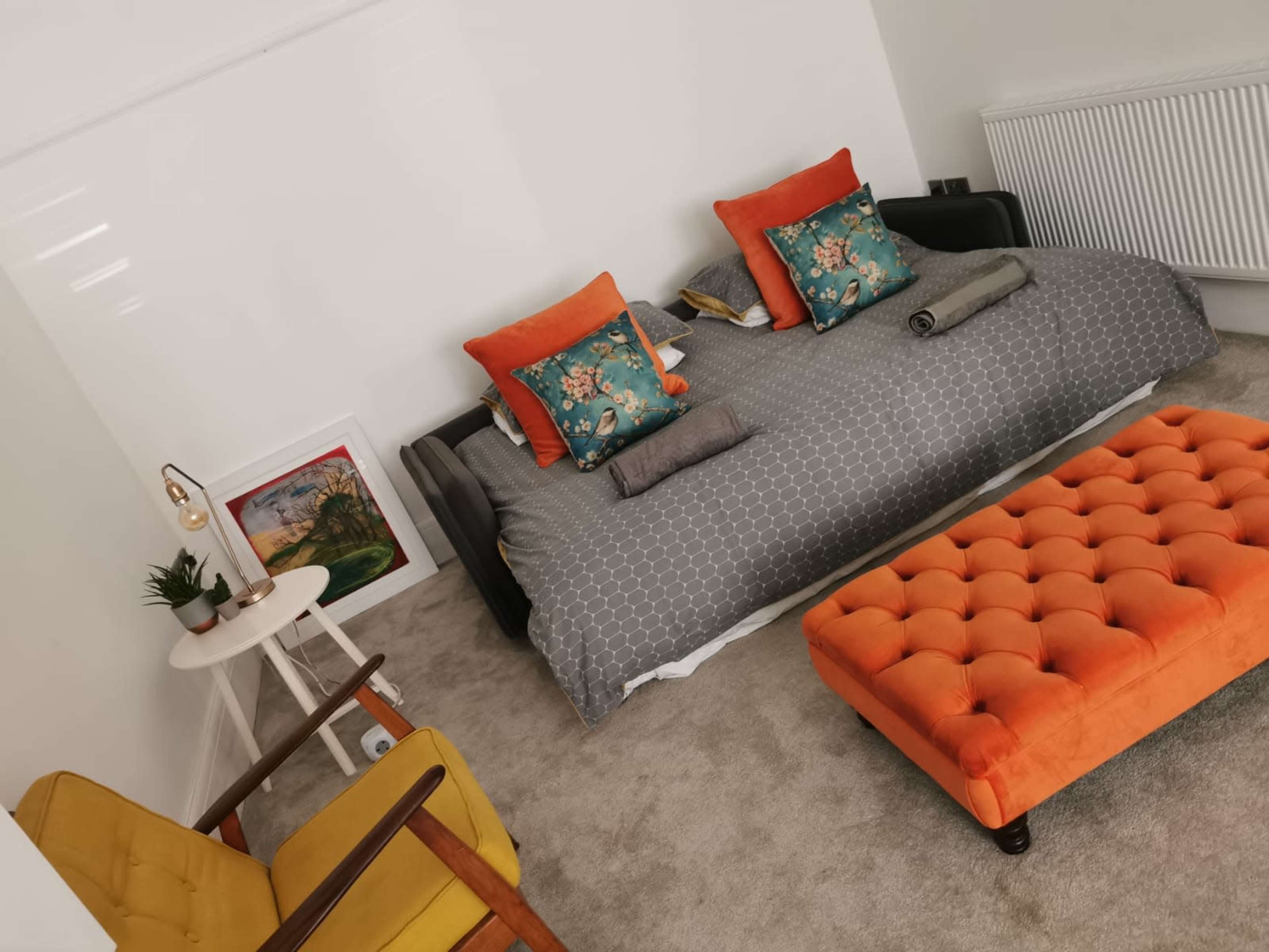 A cozy bedroom featuring a patterned bed with orange and teal cushions, two armchairs, a small table with a lamp, and an orange tufted ottoman.