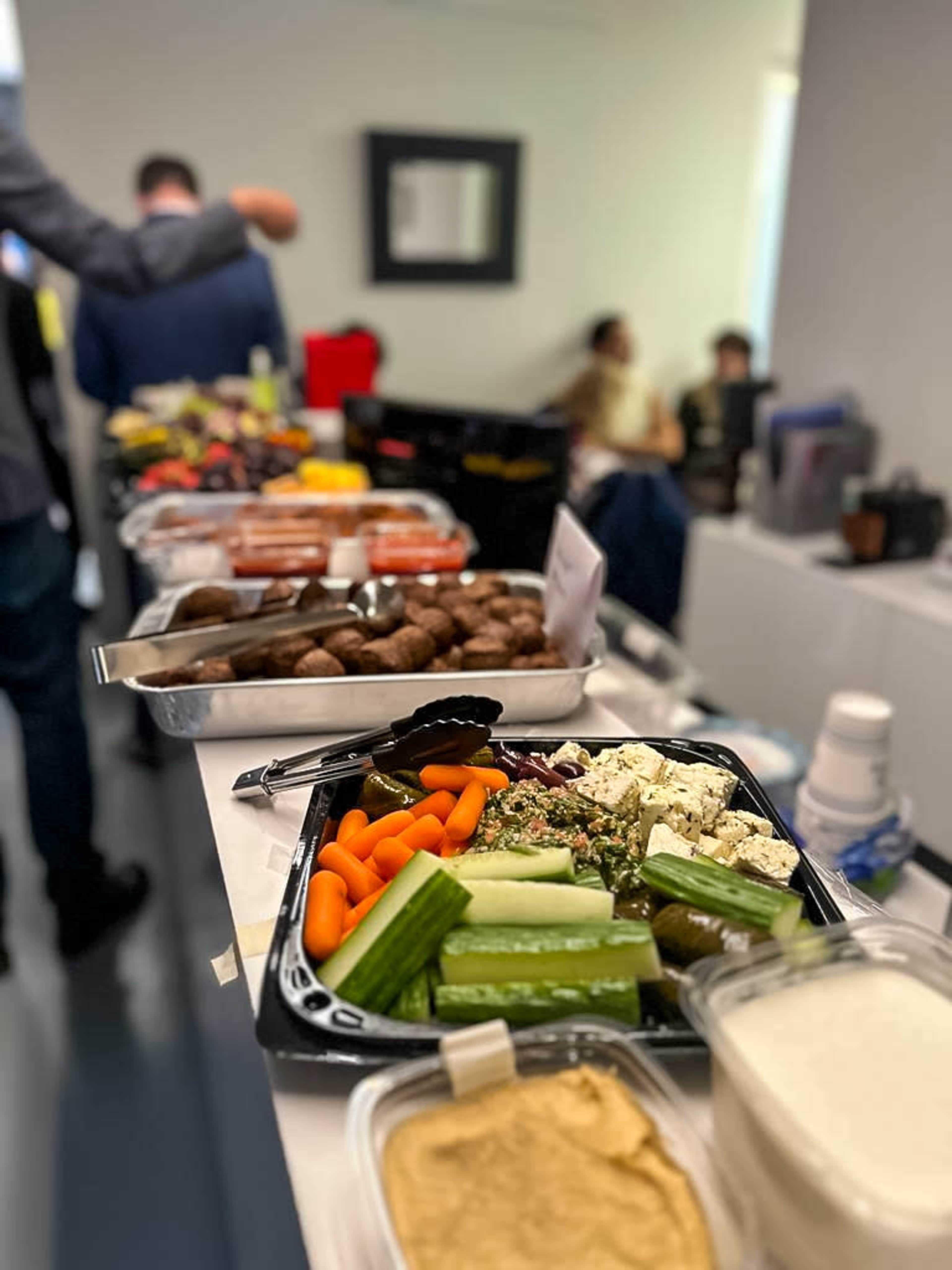 A table is set with an assortment of food items, including vegetables, hummus, and various snacks, in a brightly lit room with people in the background.