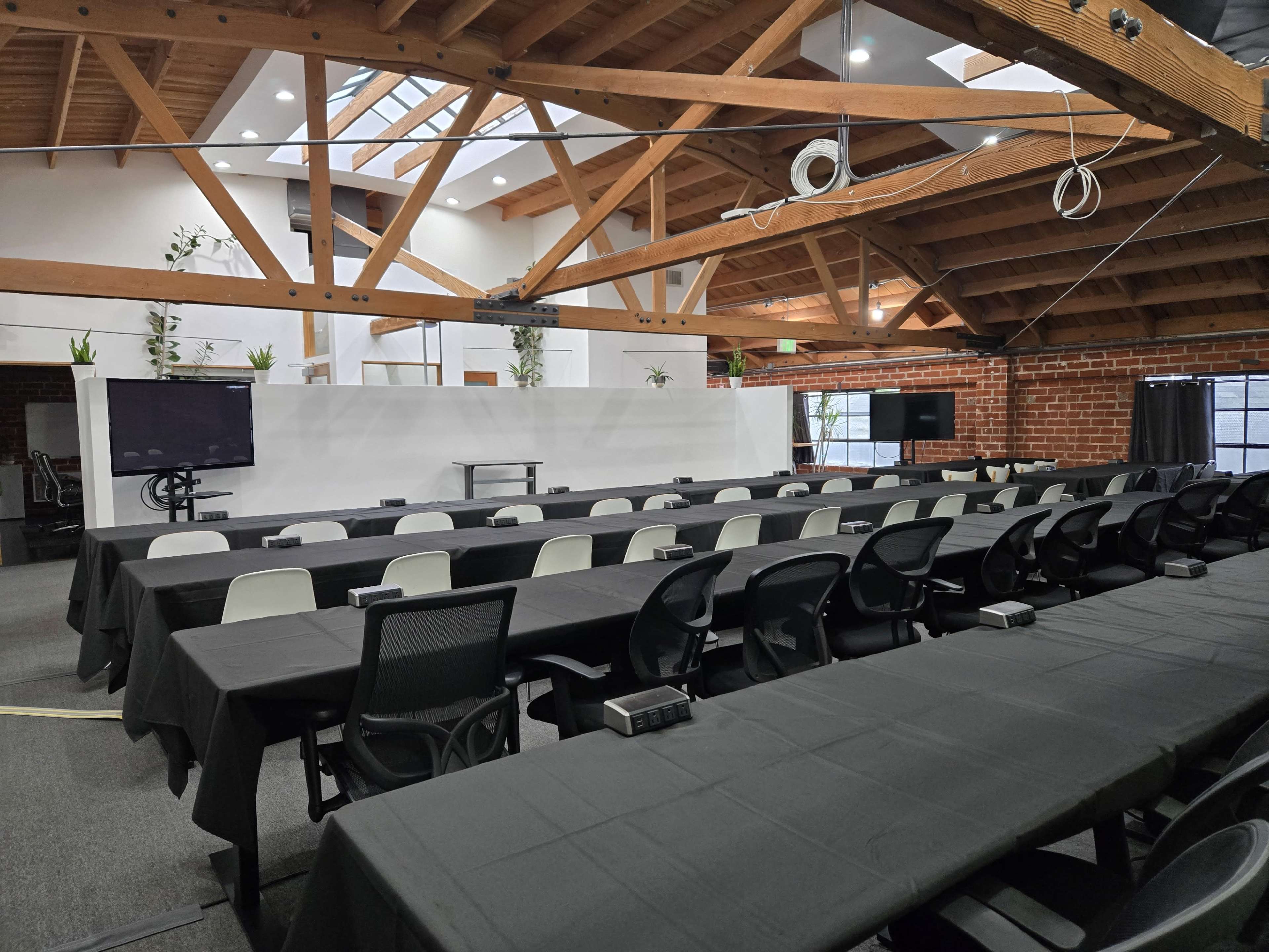 The image shows a spacious conference room with long tables covered in black cloth, arranged in rows and equipped with technology, beneath a wooden beam ceiling.