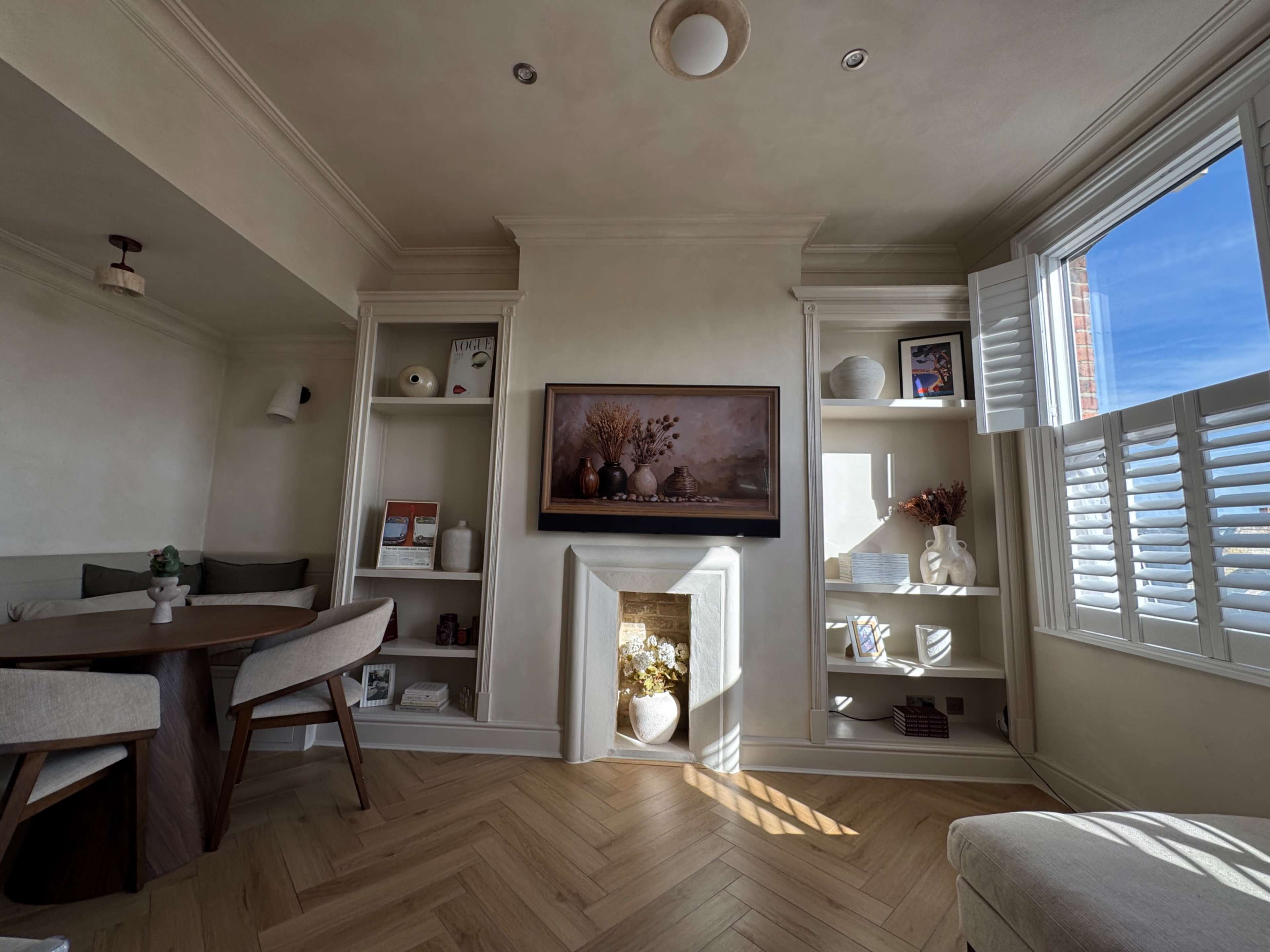 A cozy living room features a circular dining table, built-in shelves with decorative items, and a fireplace under a framed artwork, with natural light streaming through the window.