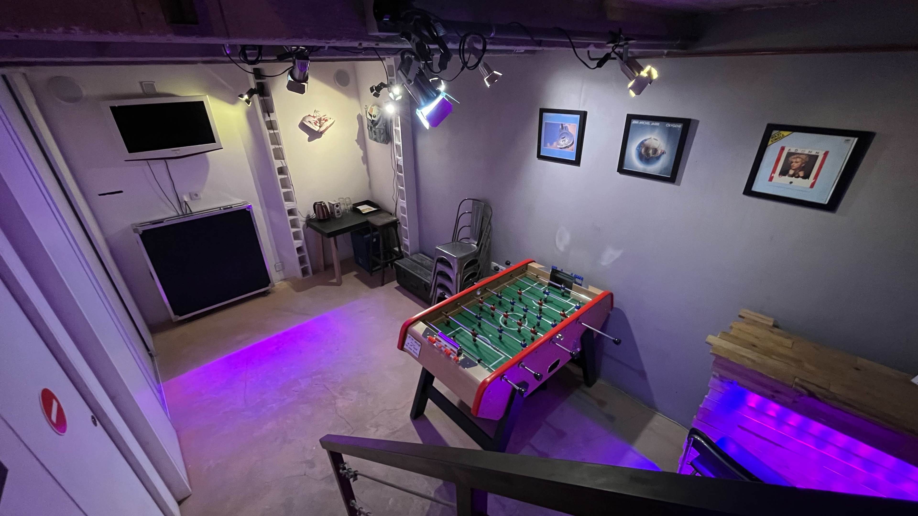 The image shows a dimly lit game room with a foosball table, framed pictures on the walls, and a small kitchenette in the background.
