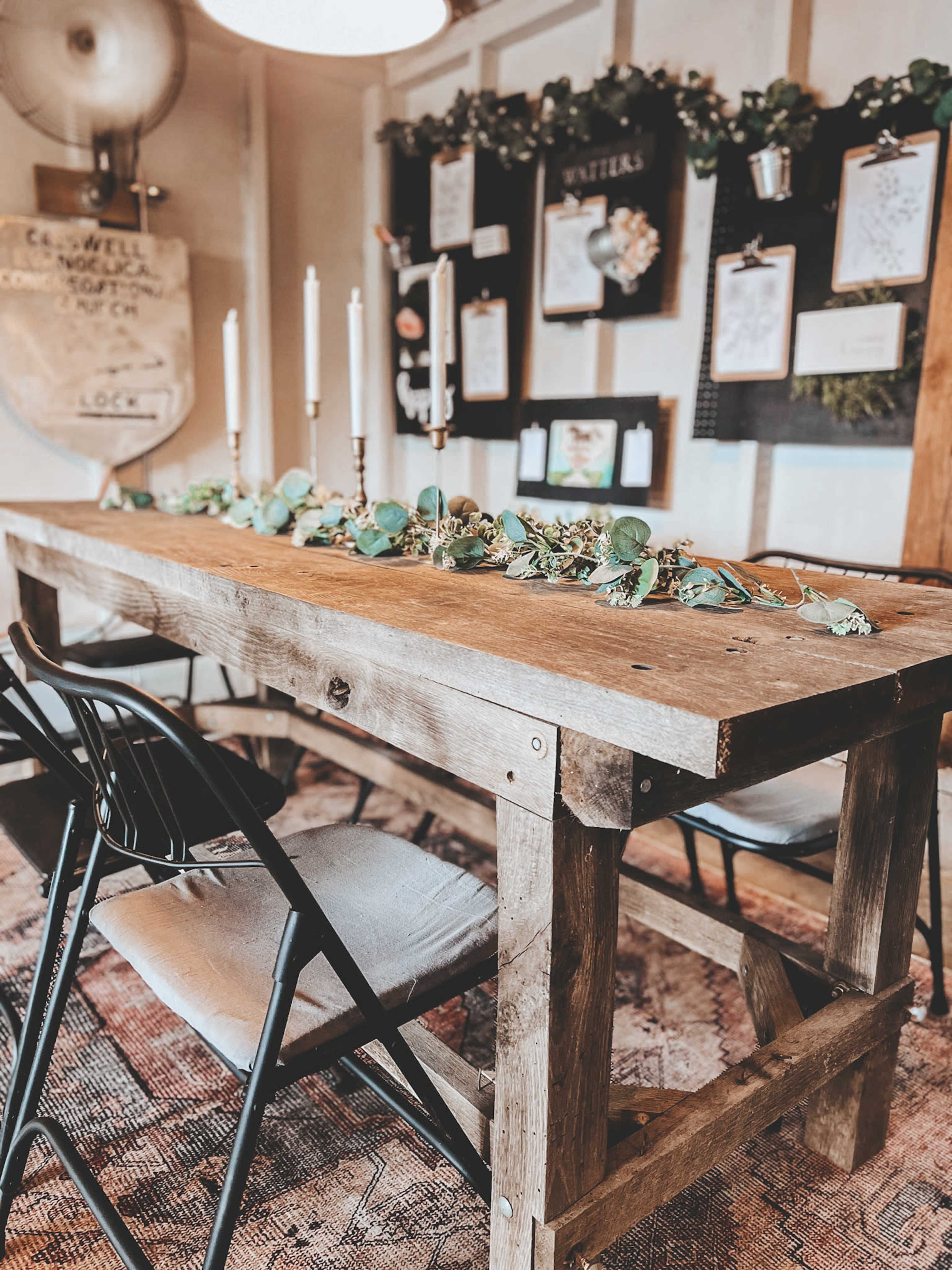 A rustic wooden table is set with candles and adorned with greenery, surrounded by black chairs in a decorated room featuring presentation boards.
