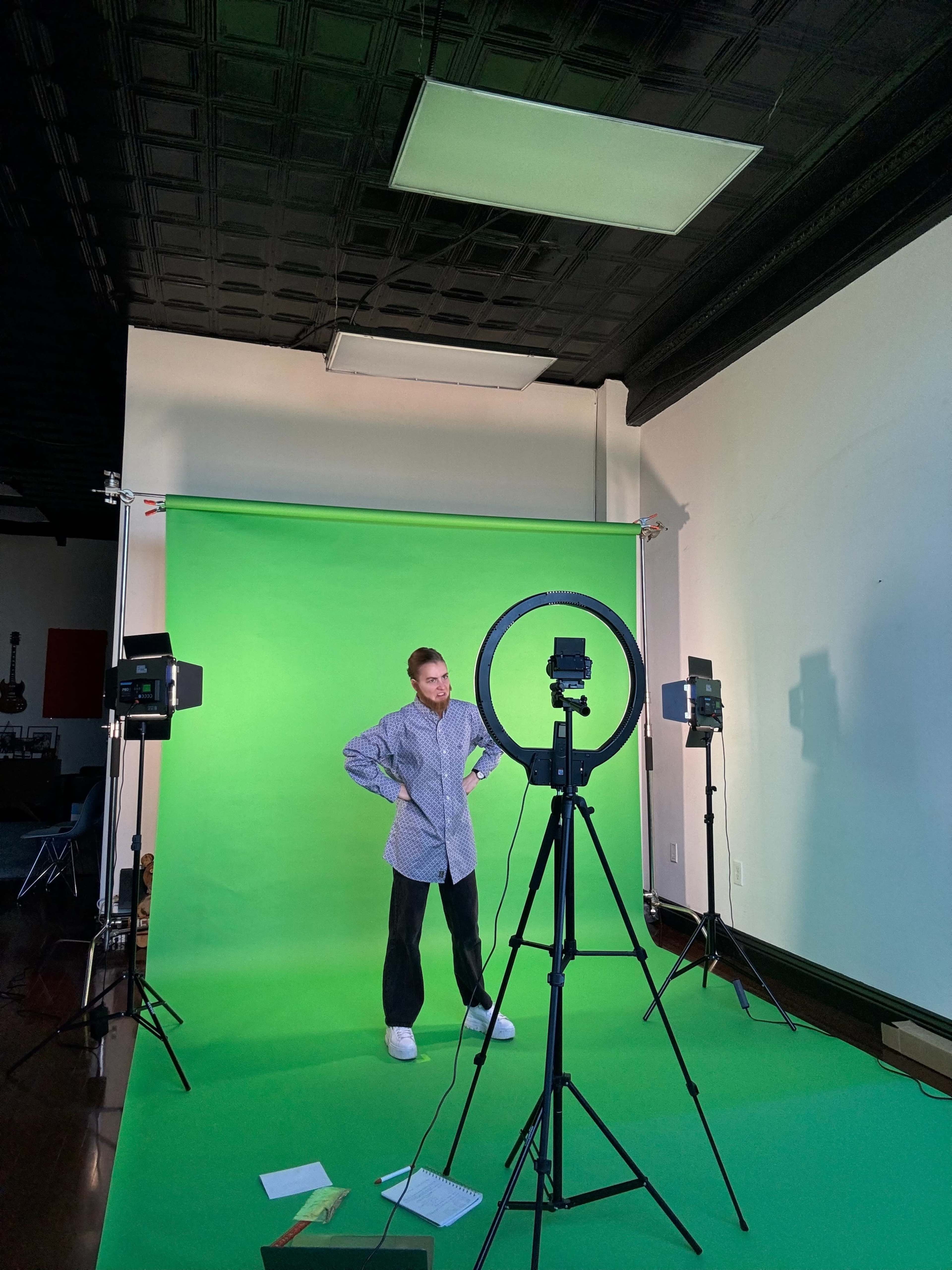A person stands in front of a green screen setup, flanked by lights and a ring light.