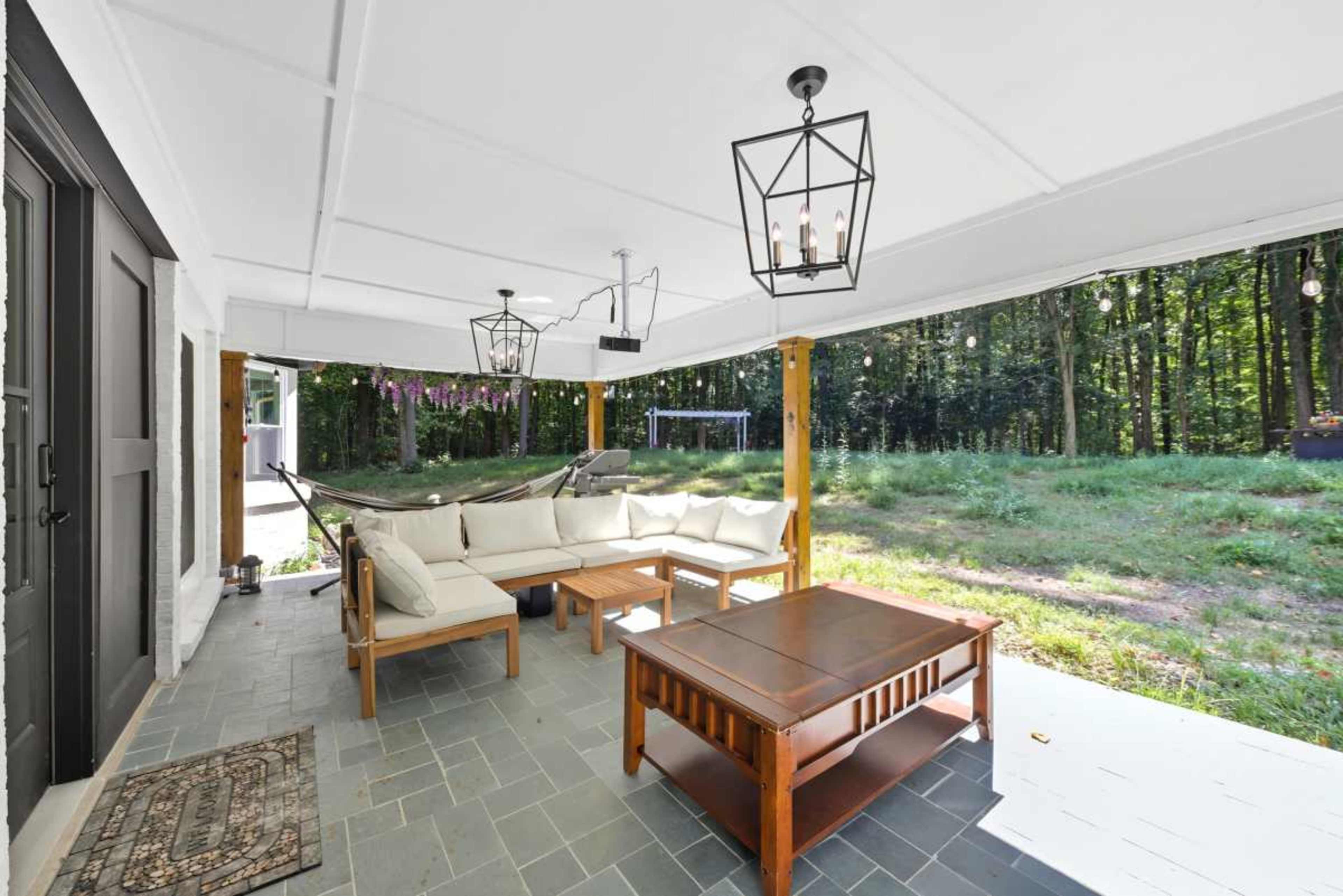 The image shows a covered patio with a sectional sofa, a coffee table, and pendant lighting, surrounded by a grassy area and trees.