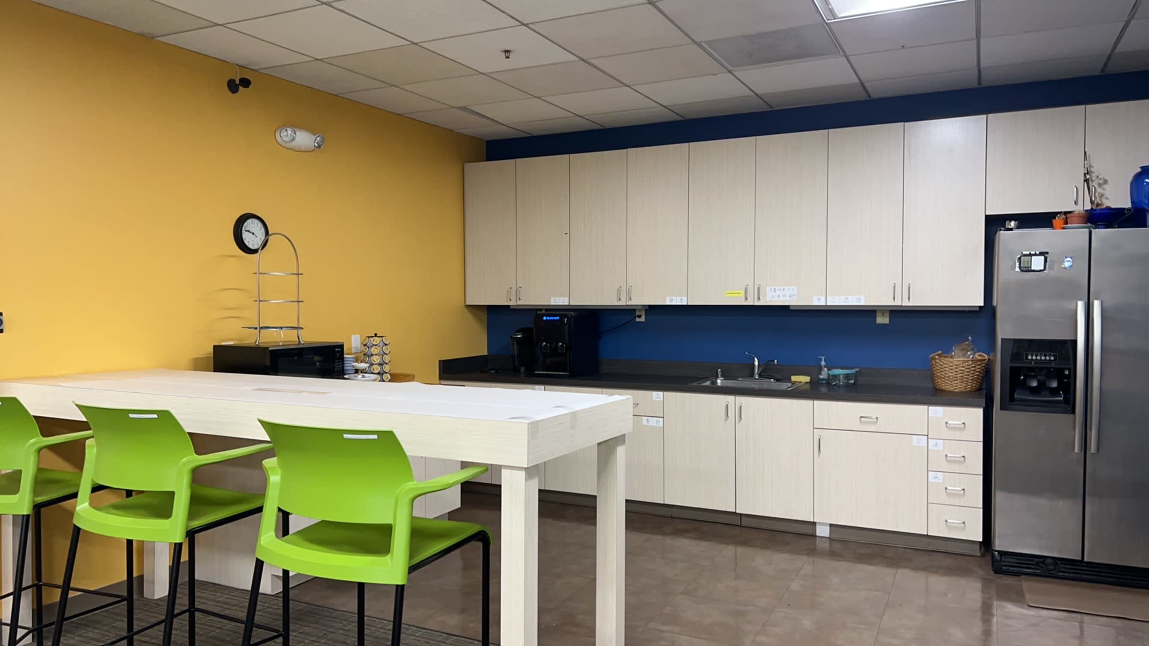 A modern kitchen with light-colored cabinets, a dark countertop, a clock on the wall, and green chairs around a tall table.