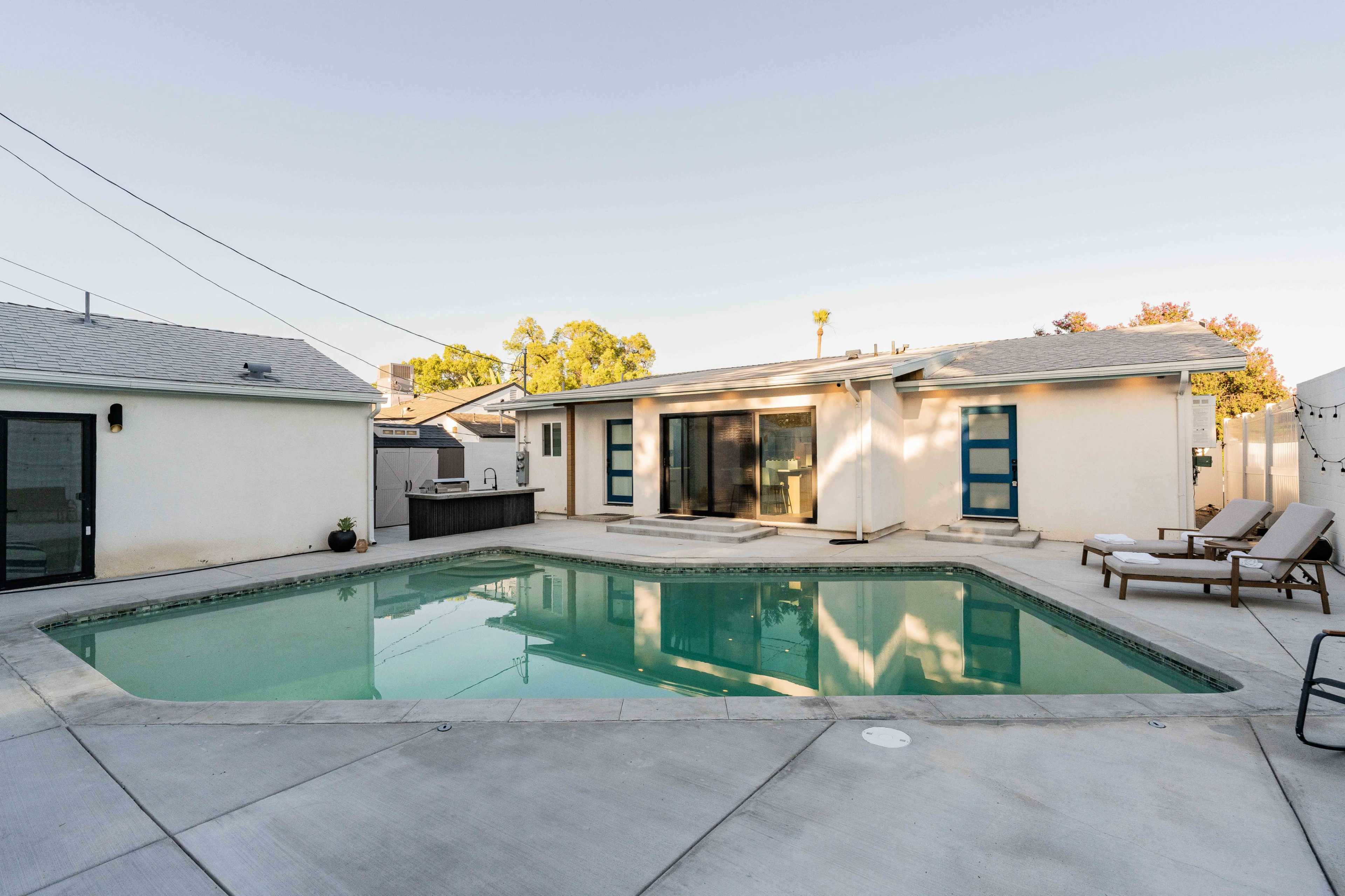 A rectangular swimming pool is surrounded by a concrete patio, with a single-story house featuring large glass doors on one side.