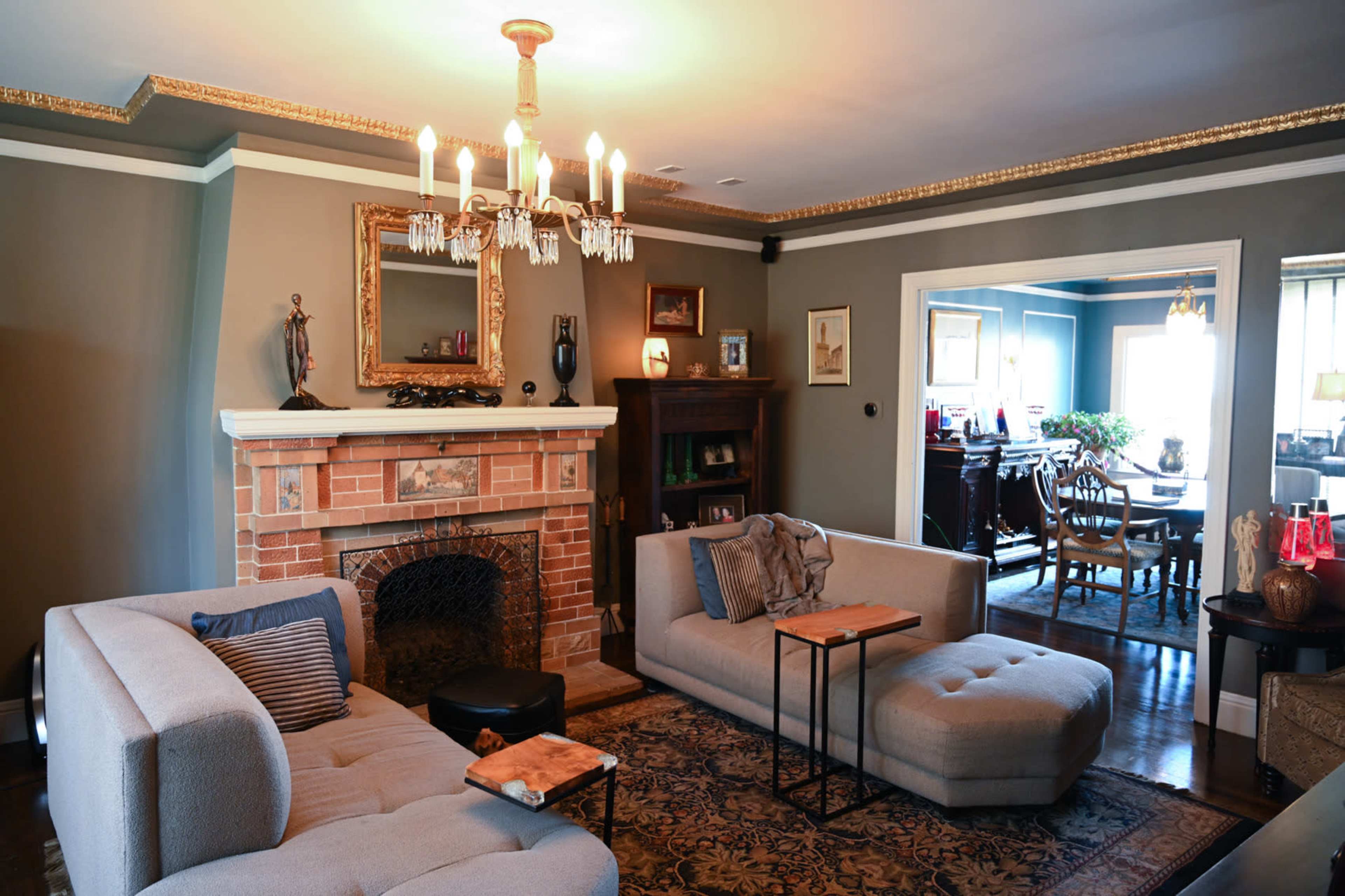 A living room features a brick fireplace, two gray sofas, and a chandelier, with a dining area visible through an open doorway.