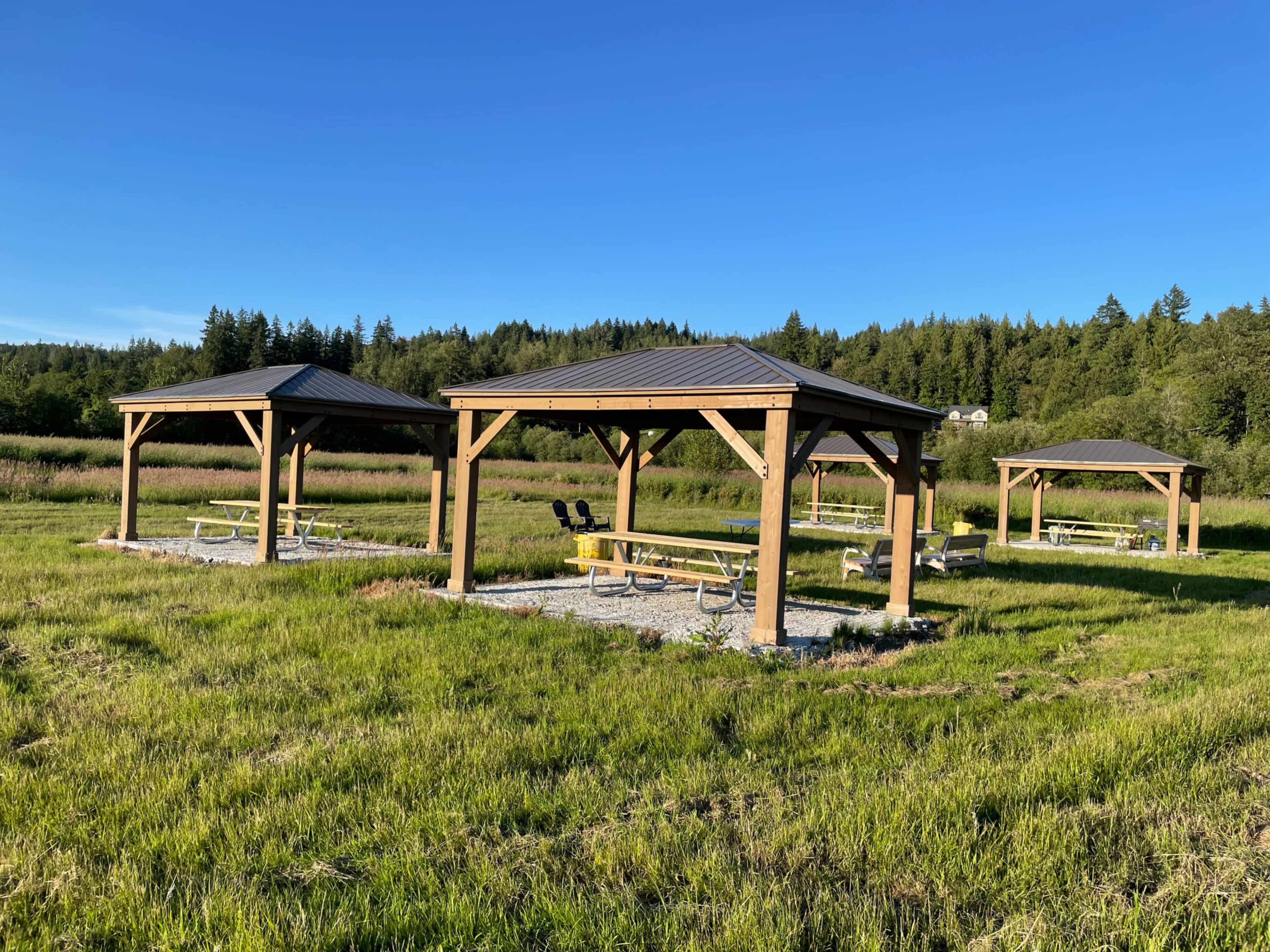 Three wooden pavilions with metal roofs set in a grassy field surrounded by trees.