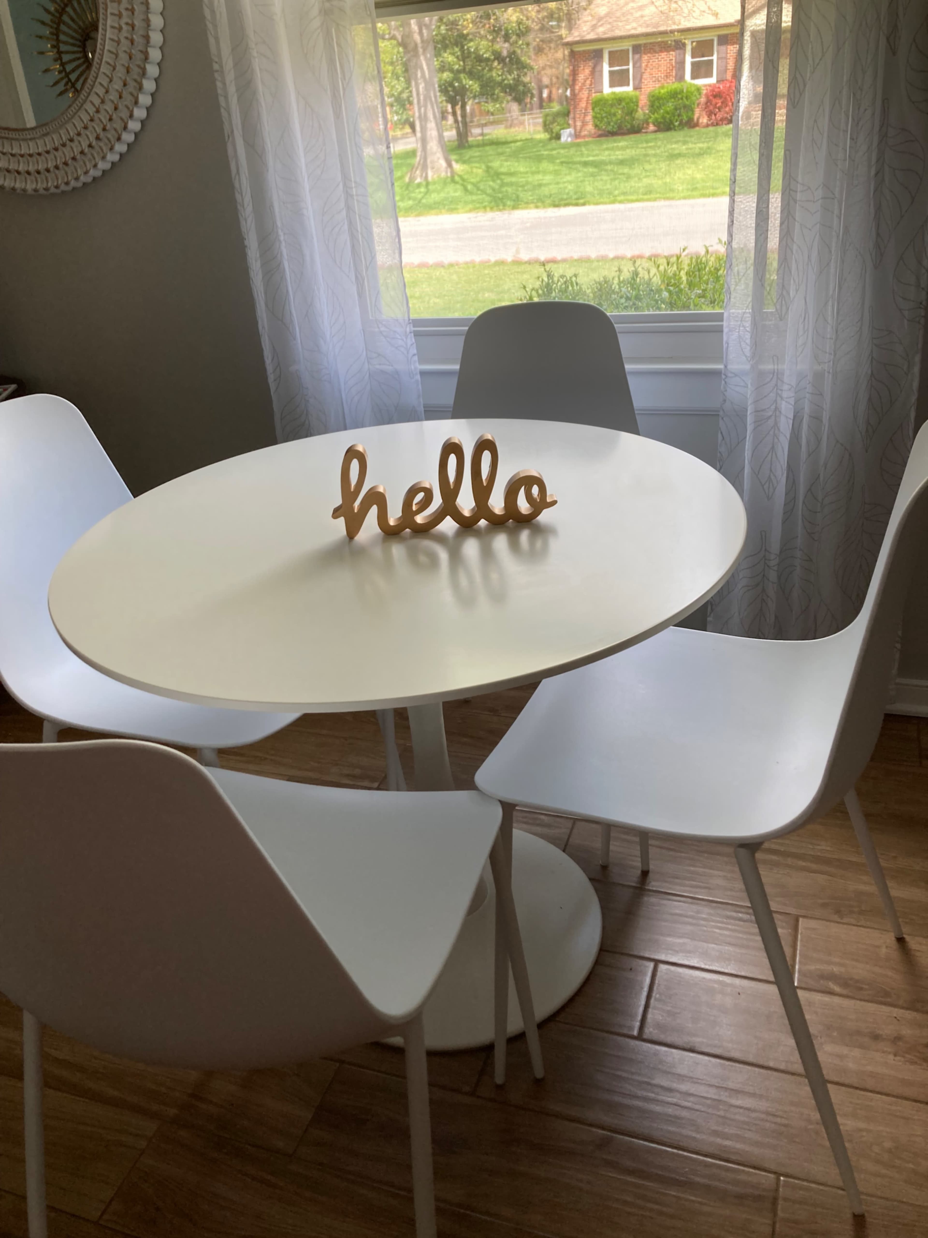 A round white table with four white chairs is set near a window, featuring a decorative wooden "hello" sign in the center.