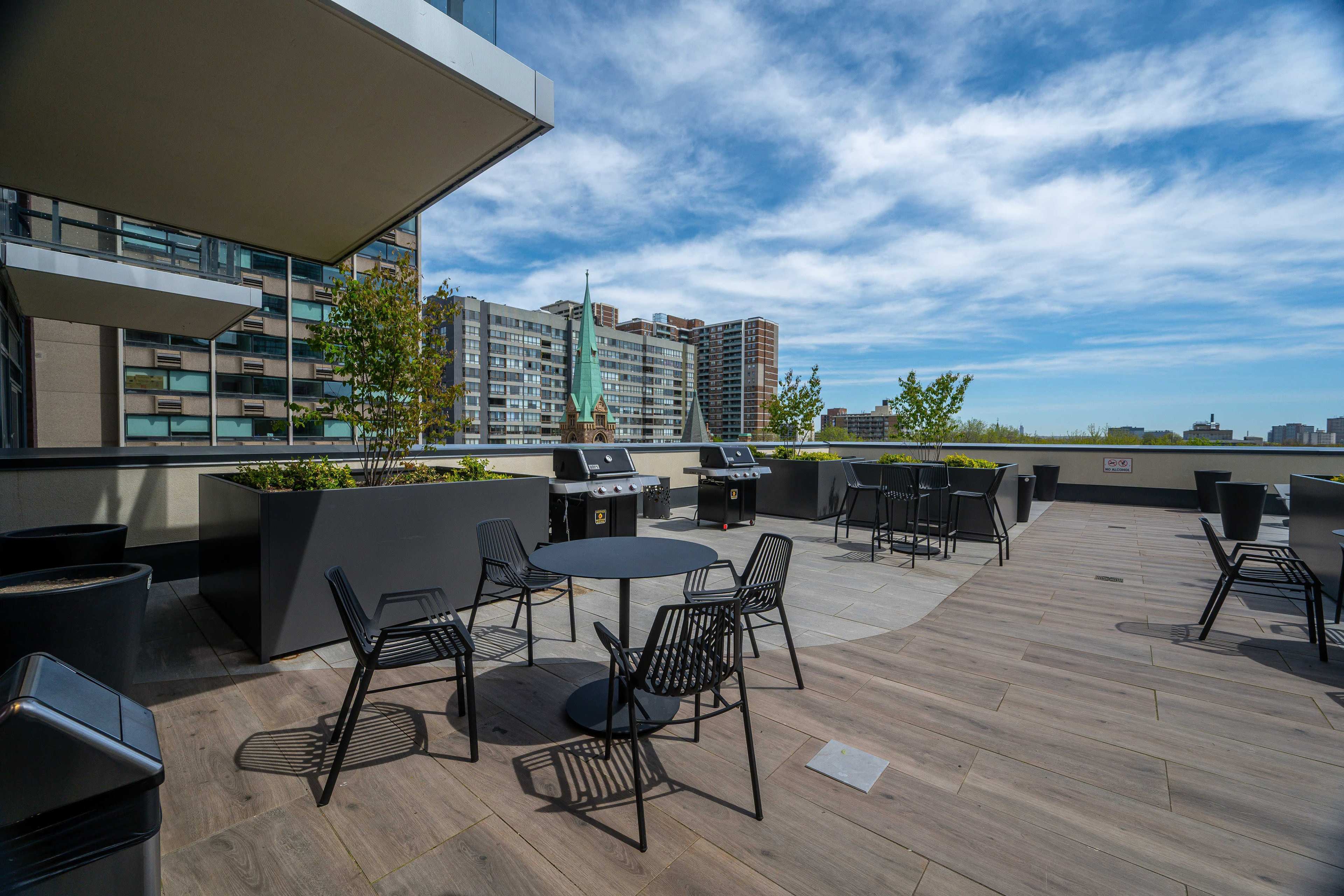 The image shows a rooftop patio with black metal chairs and tables, surrounded by planters, and a skyline of buildings under a blue sky with scattered clouds.