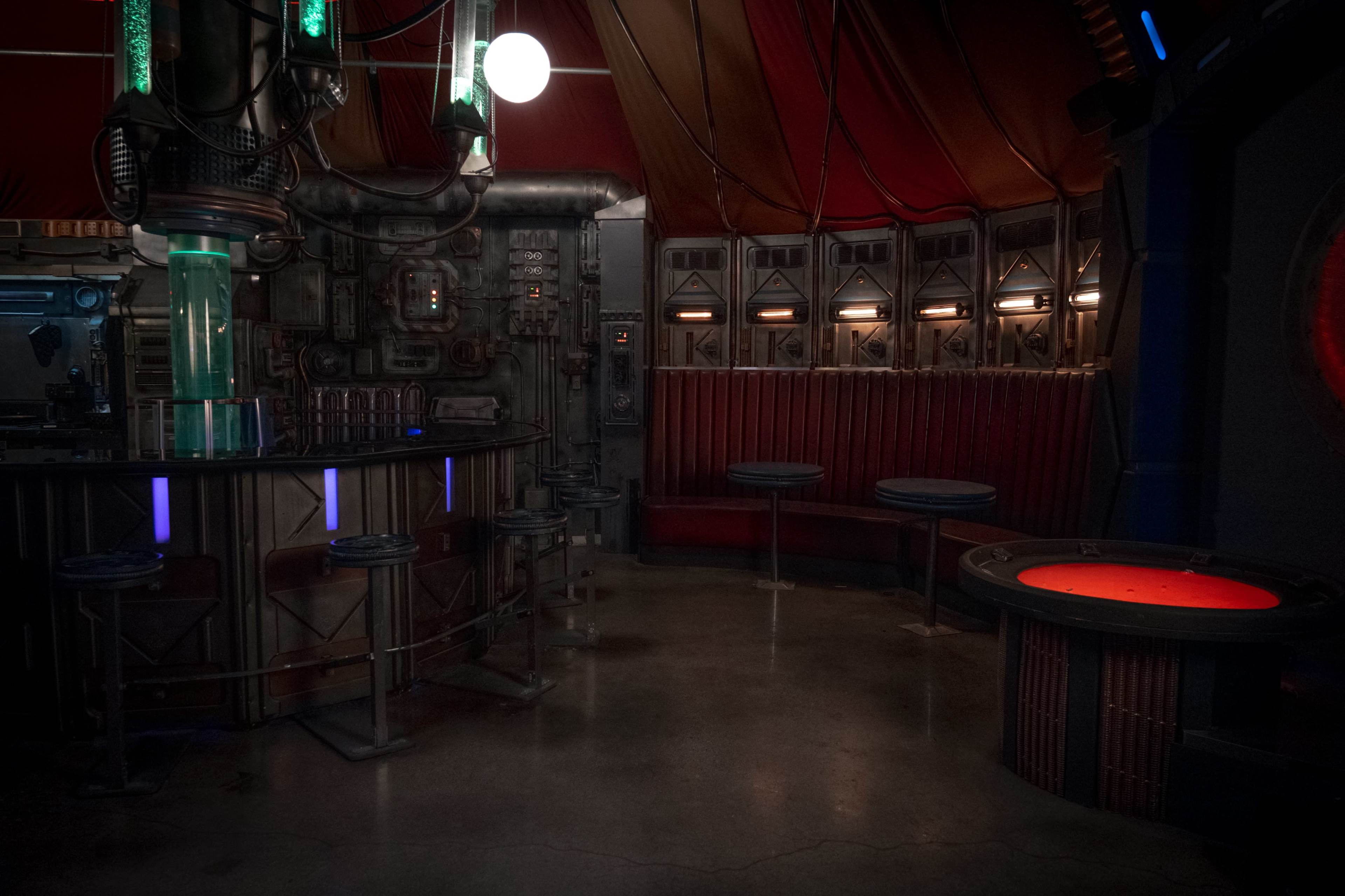 The image shows a dimly lit, futuristic bar area with metallic fixtures and round seating arranged around a circular table.