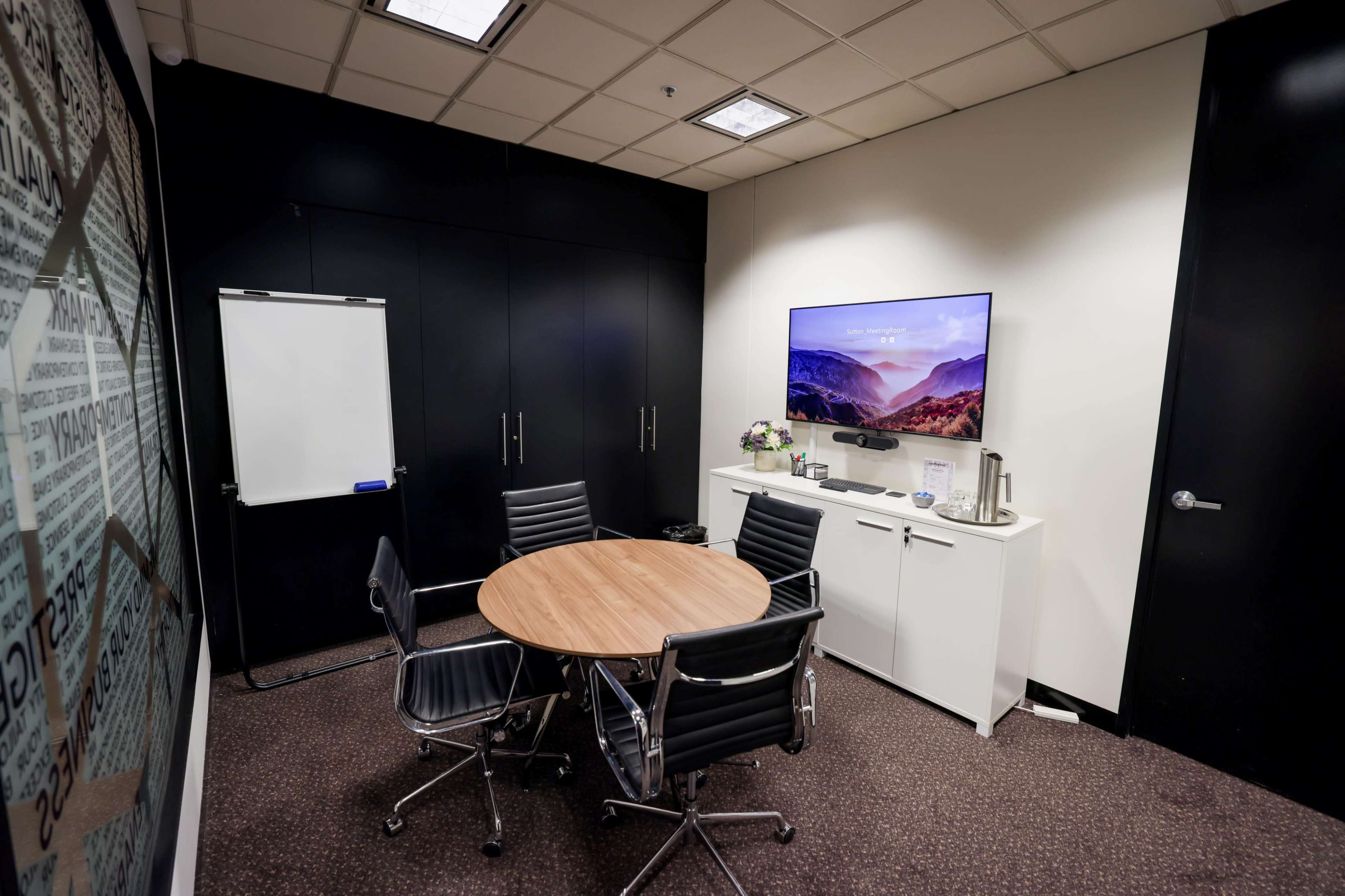 A small meeting room features a circular wooden table surrounded by four black chairs, a whiteboard, and a television mounted on the wall.