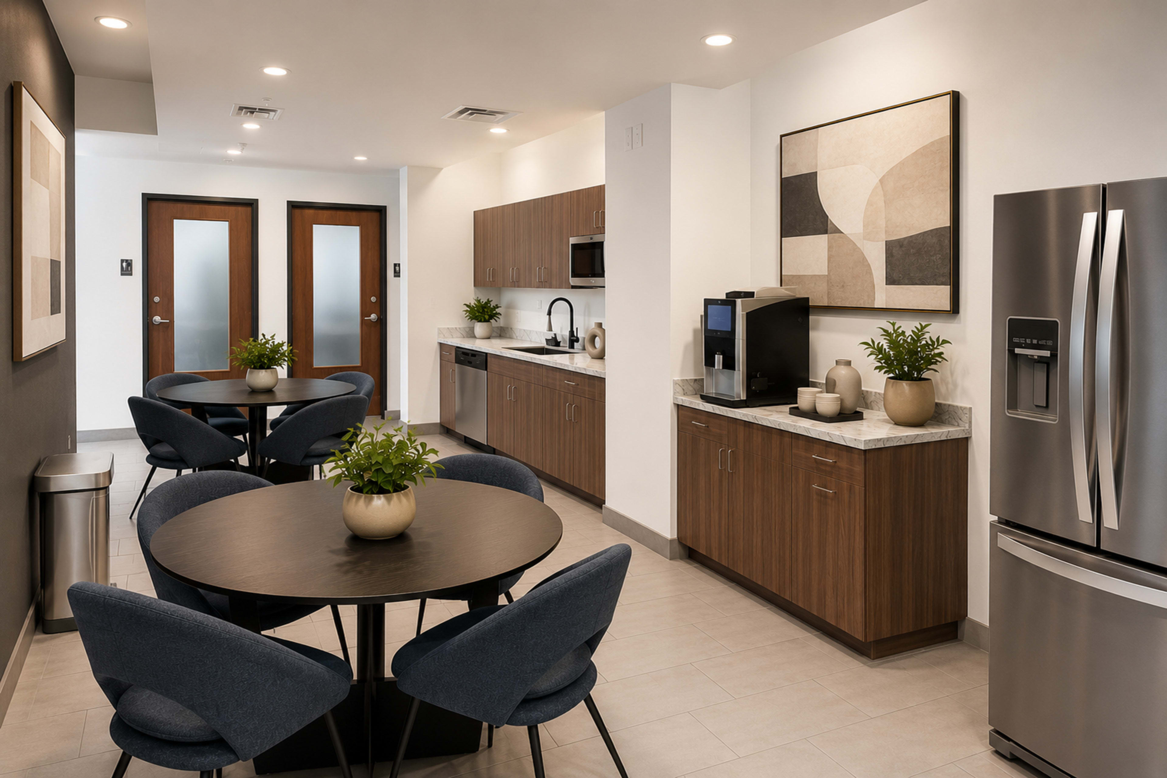 The image shows a modern kitchen area with a circular dining table and chairs, cabinets, a refrigerator, and a coffee station against a light-colored wall.
