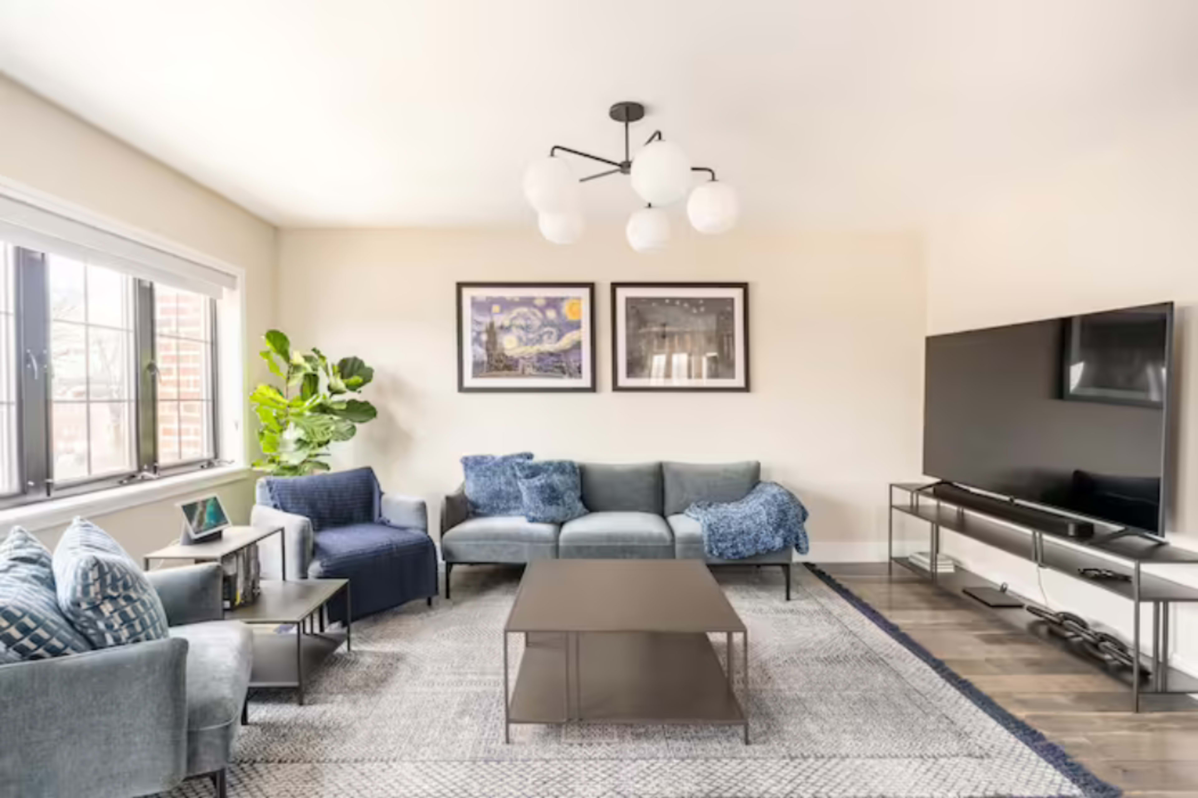 A living room with a sectional sofa, armchair, coffee table, and a large television mounted on the wall, decorated with framed artwork and a potted plant.