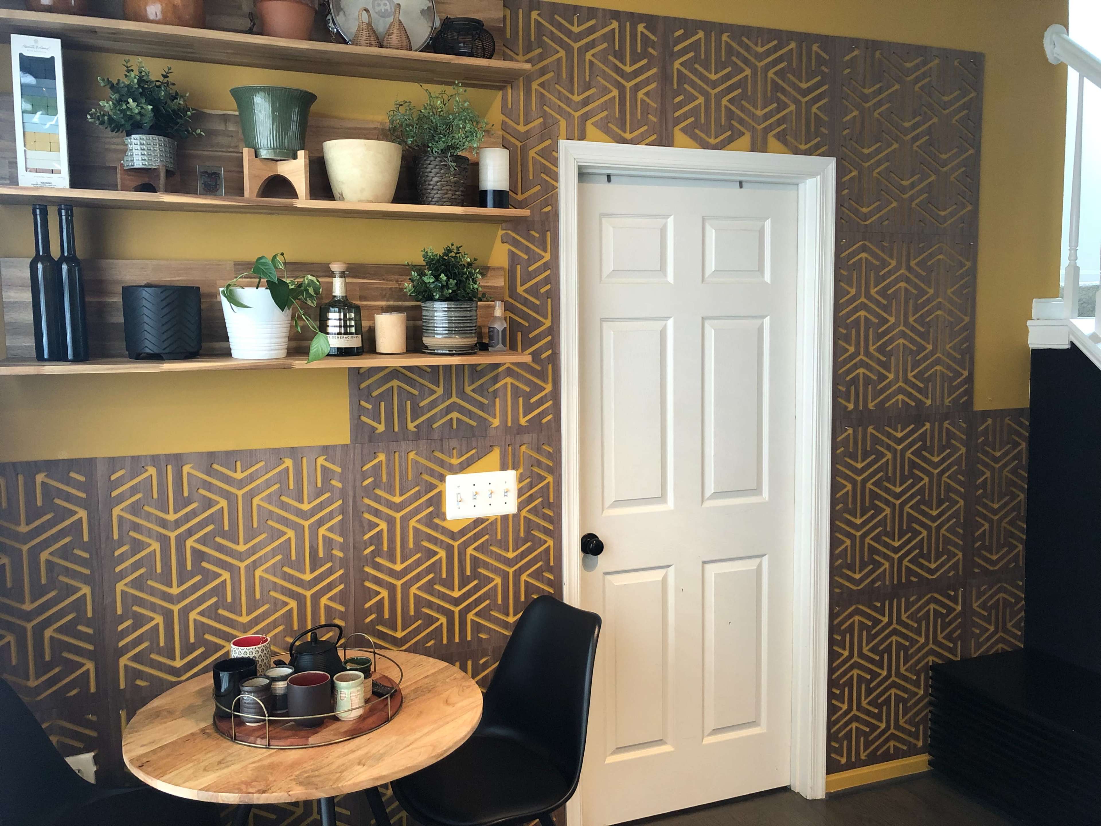 The image shows a small dining area featuring a round wooden table with chairs, shelves displaying various potted plants and kitchen items, and a white door set against a patterned wall.