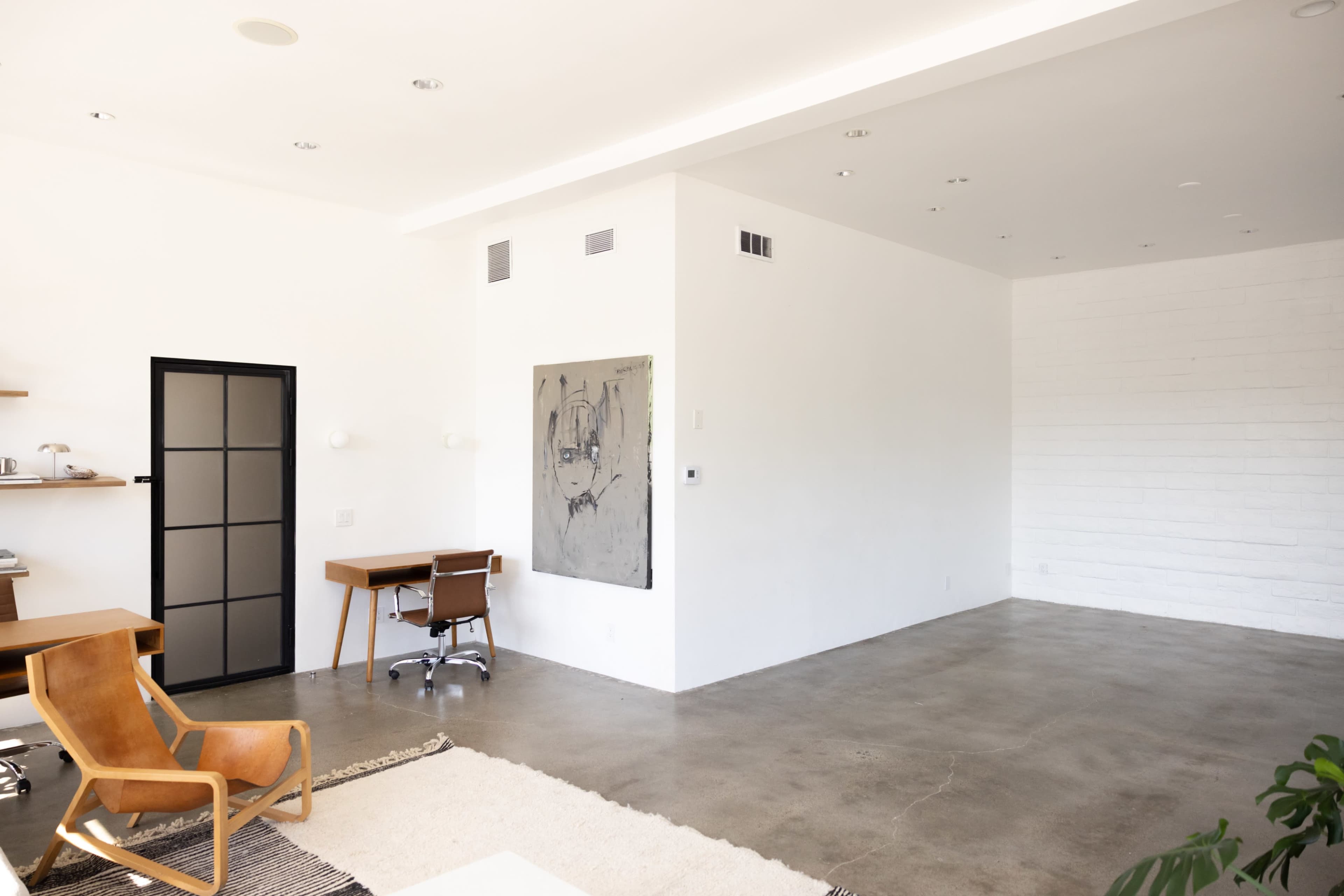 The image shows a modern, minimalistic interior space with a concrete floor, white walls, a wooden desk, and a stylish chair.