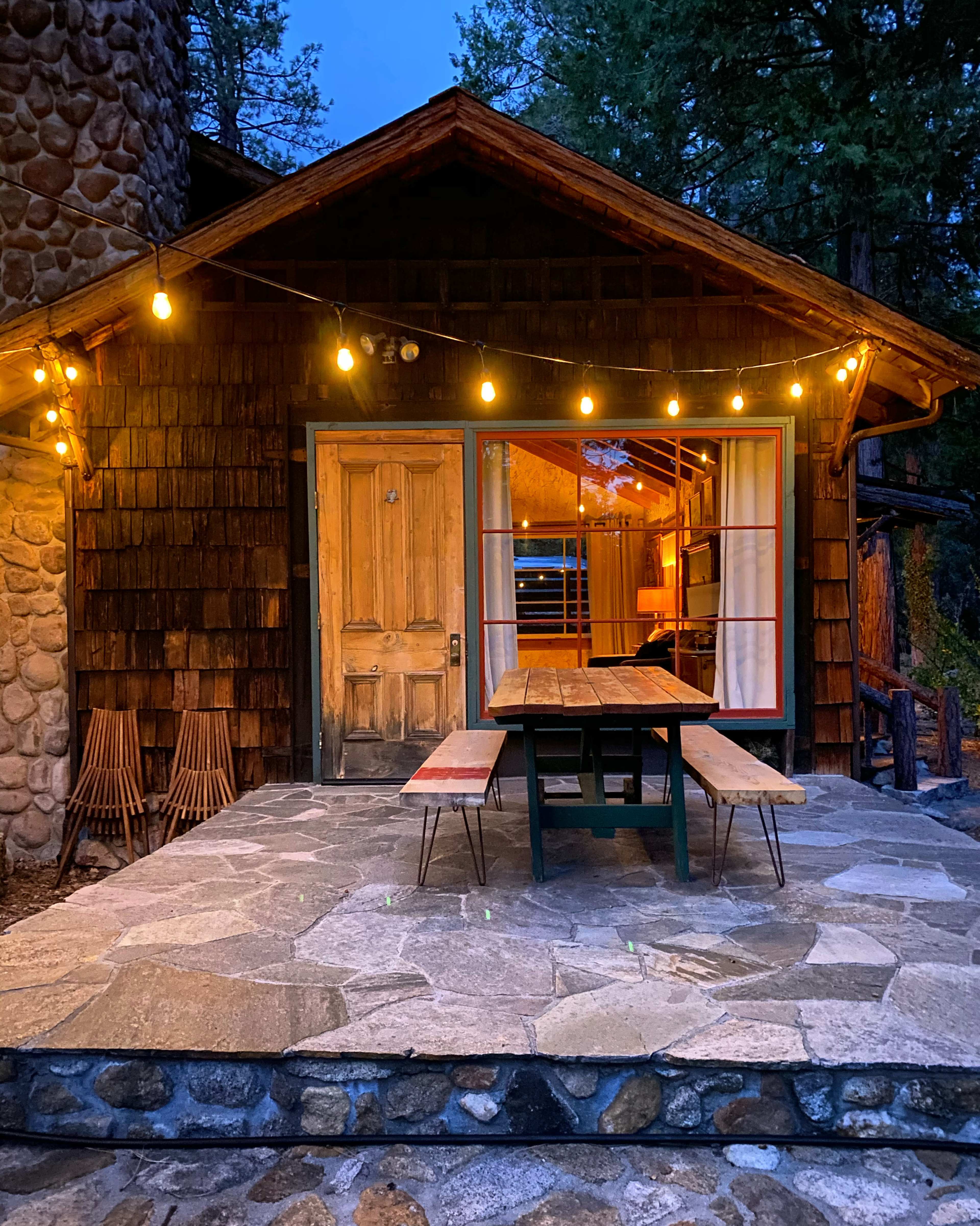 A rustic cabin with wooden siding and a stone wall features a large patio with a picnic table illuminated by string lights at dusk.