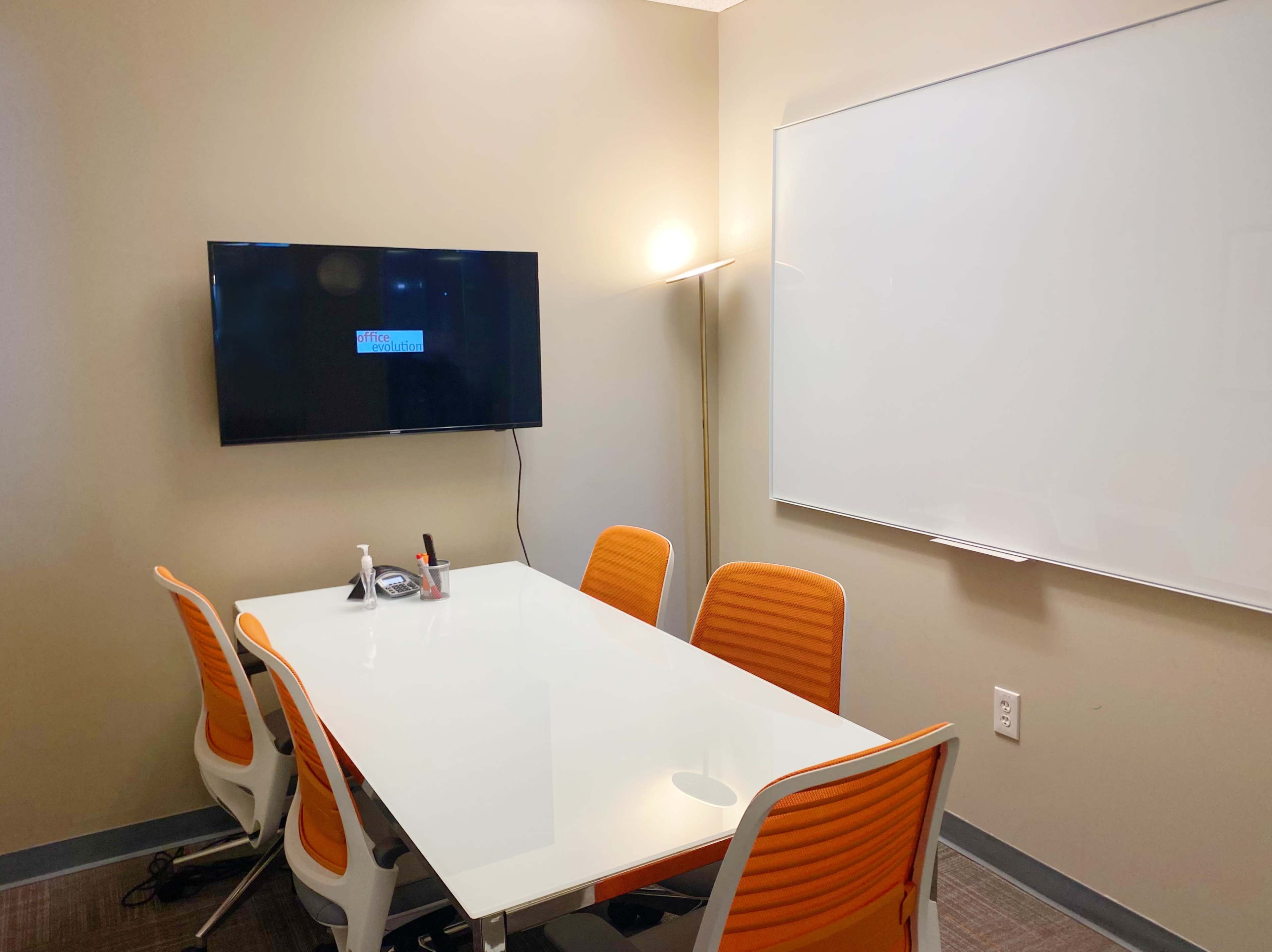 A conference room features a glass table surrounded by orange chairs, a wall-mounted television, and a whiteboard.
