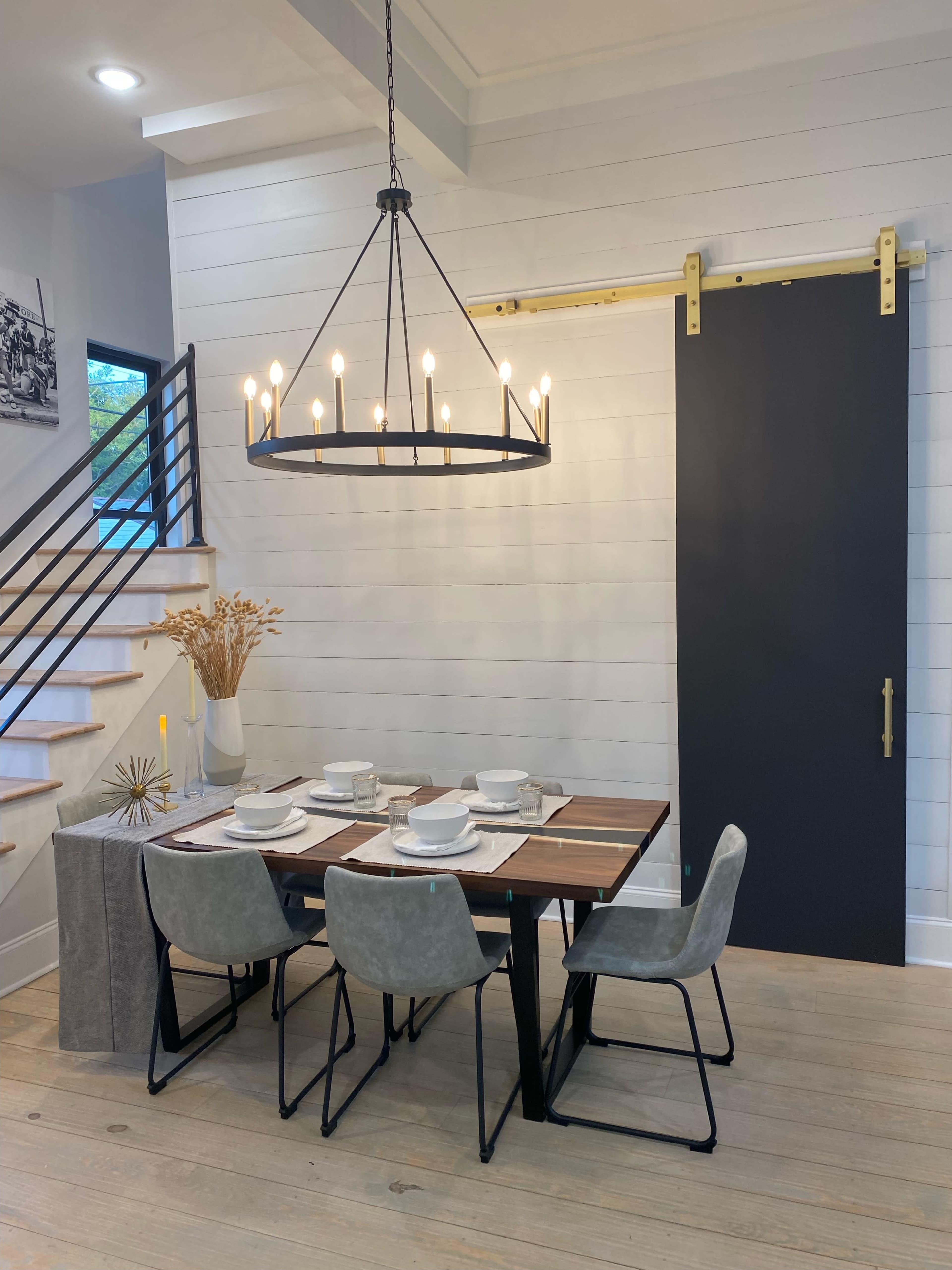 A modern dining area features a wooden table set with dishes, surrounded by four chairs, illuminated by a large chandelier, and complemented by a sliding barn door and decorative elements.