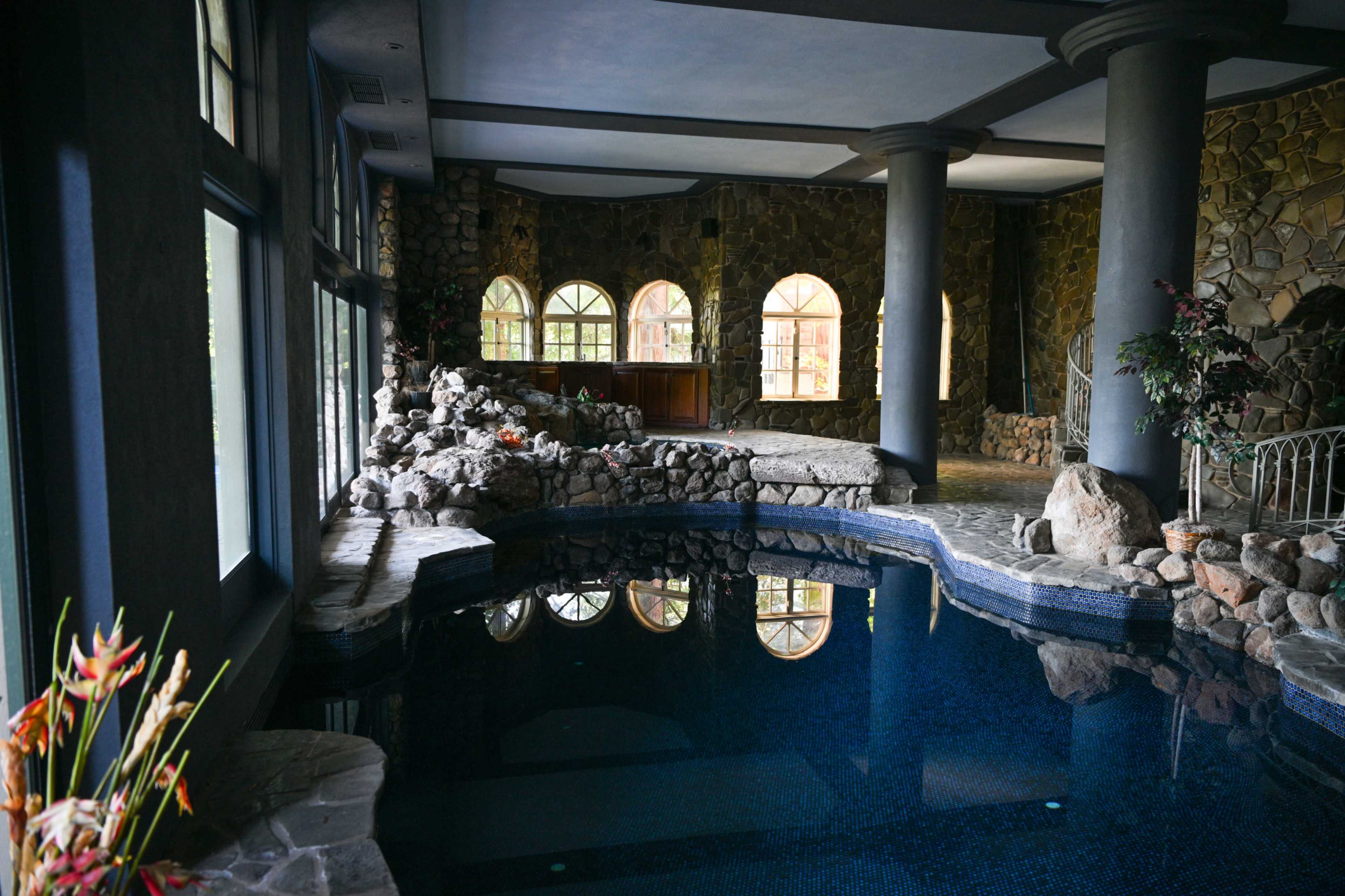 The image depicts an indoor pool area surrounded by stone walls and large windows, with a rock formation and plants adding to the natural aesthetic.