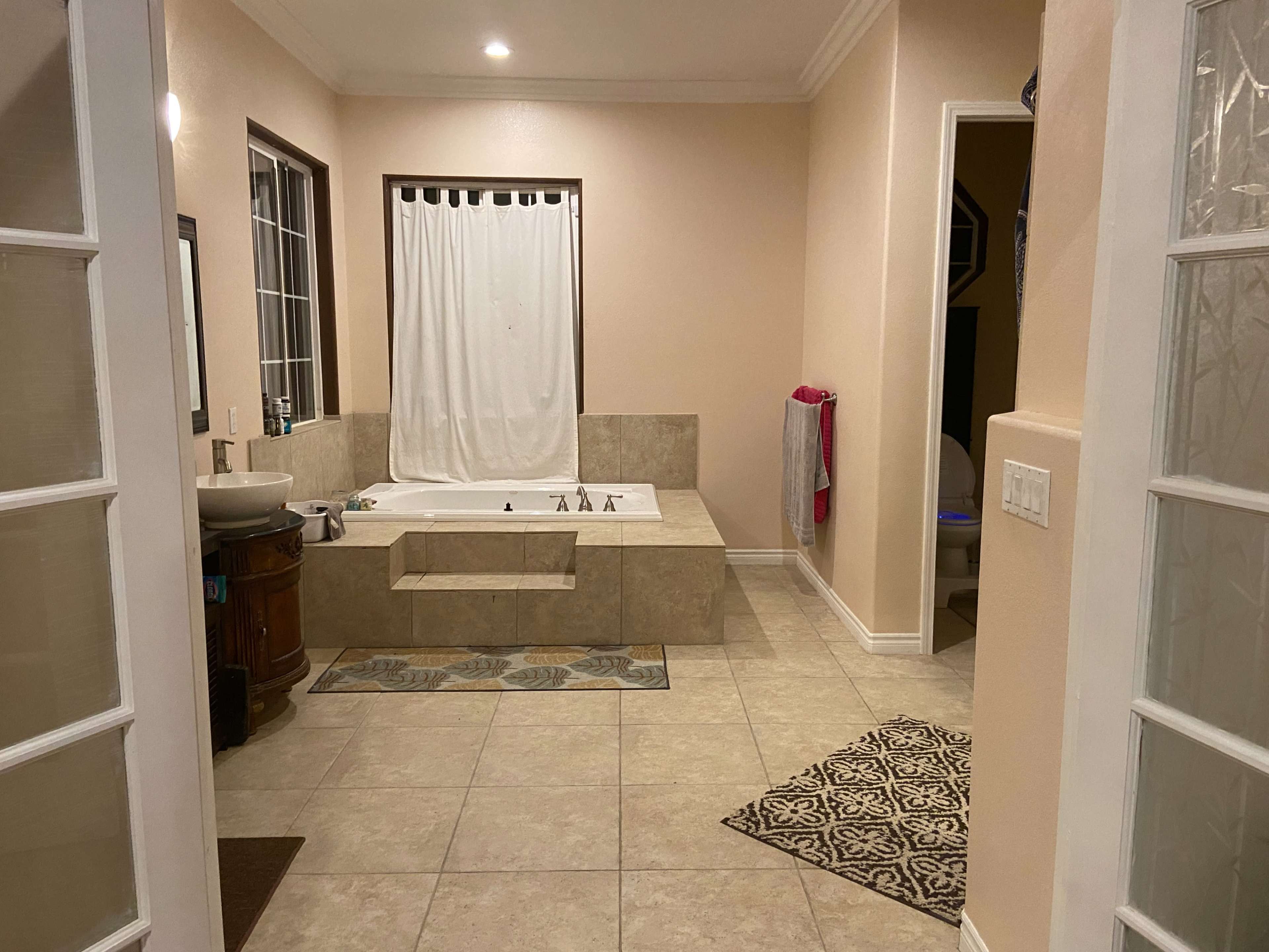 The image shows a spacious bathroom with a large bathtub, a sink, and tiled flooring under soft lighting.