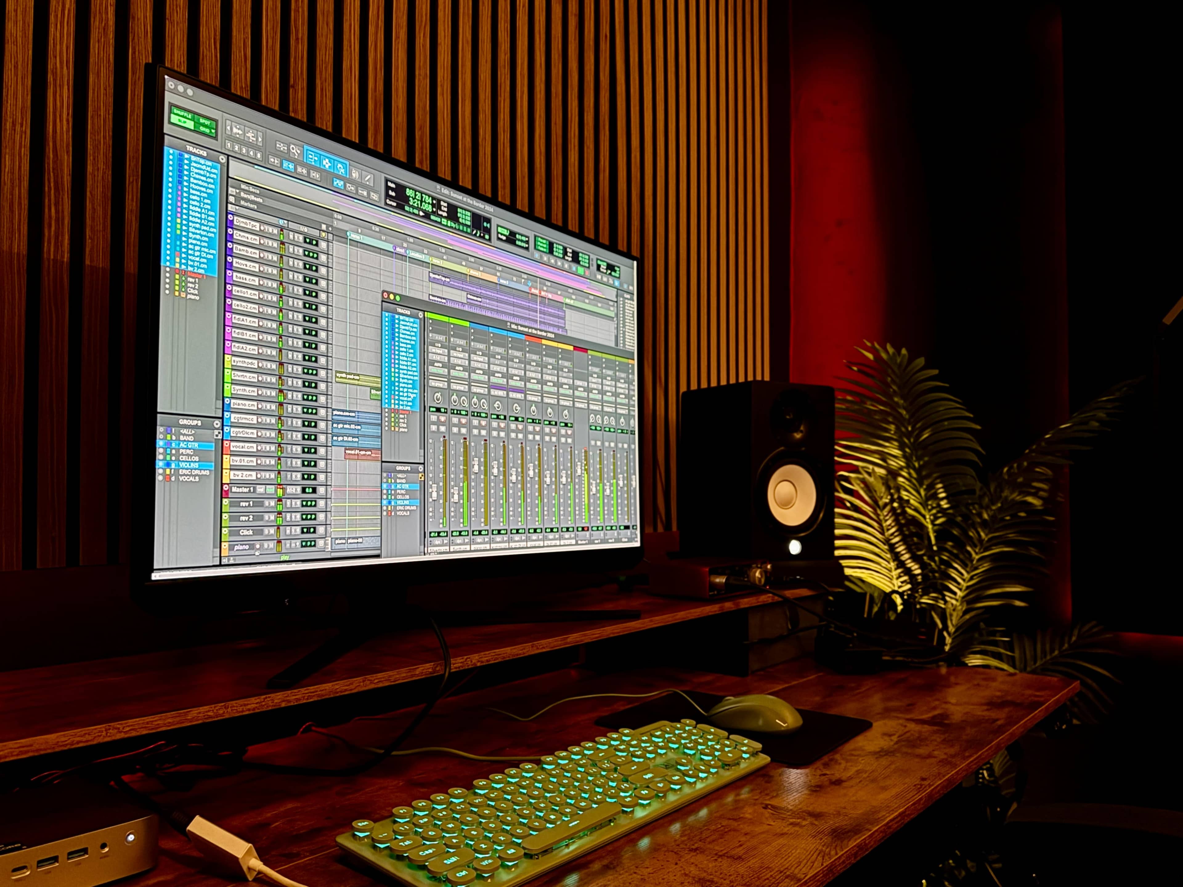 A computer screen displaying a digital audio workstation software is positioned next to a studio monitor speaker and a decorative plant on a wooden desk.