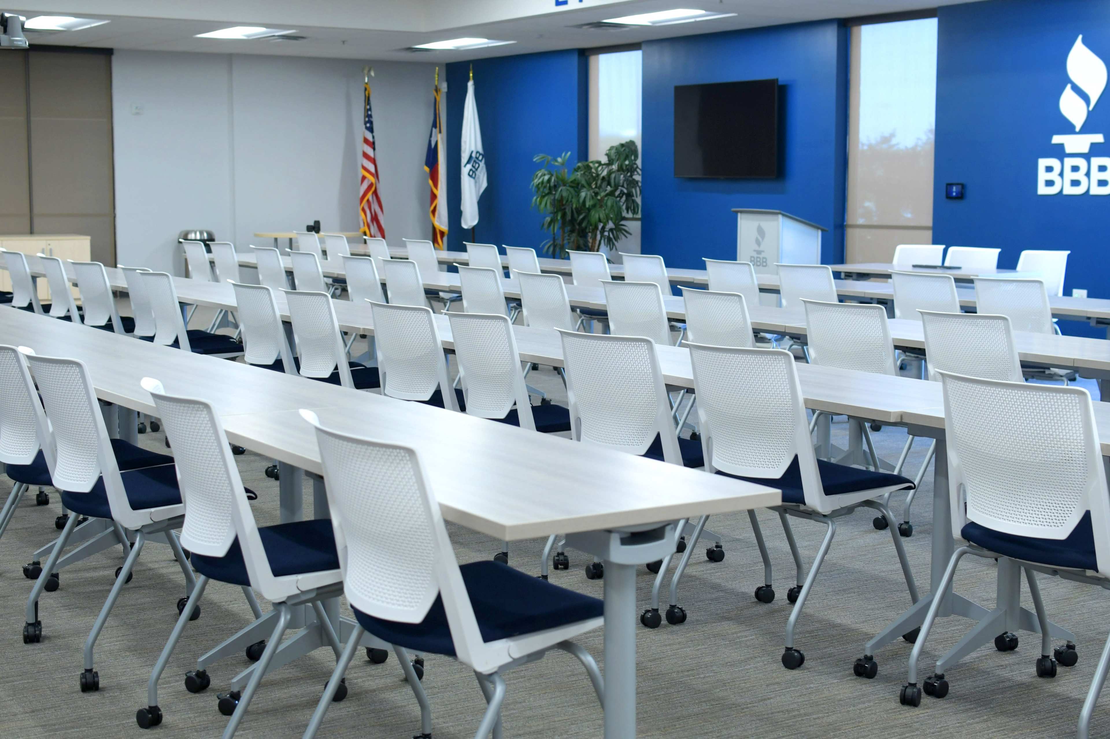 A conference room with multiple rows of tables and chairs arranged in a neat layout, featuring flags and a large screen on the wall.